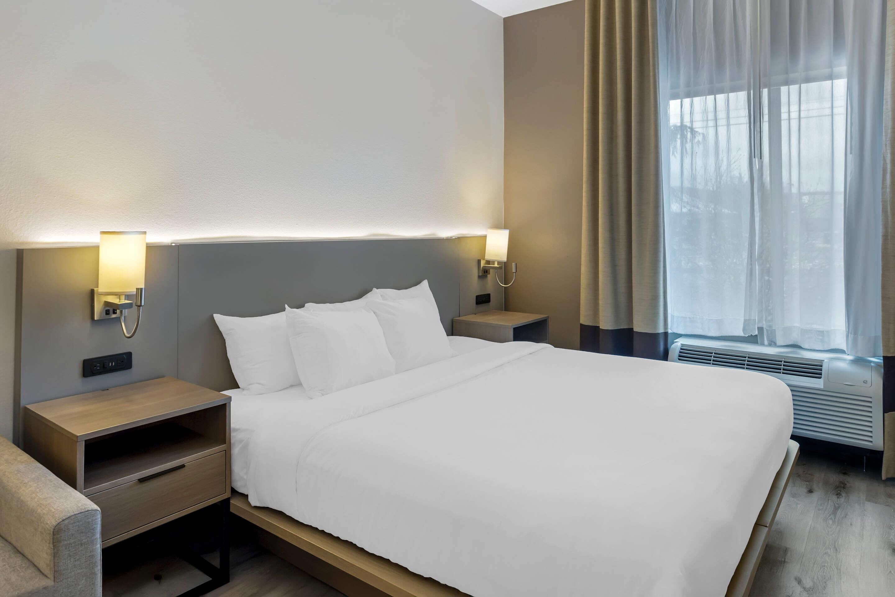 A neatly made bed with white linens, two bedside tables with lamps, a wooden nightstand, and a window with sheer curtains.