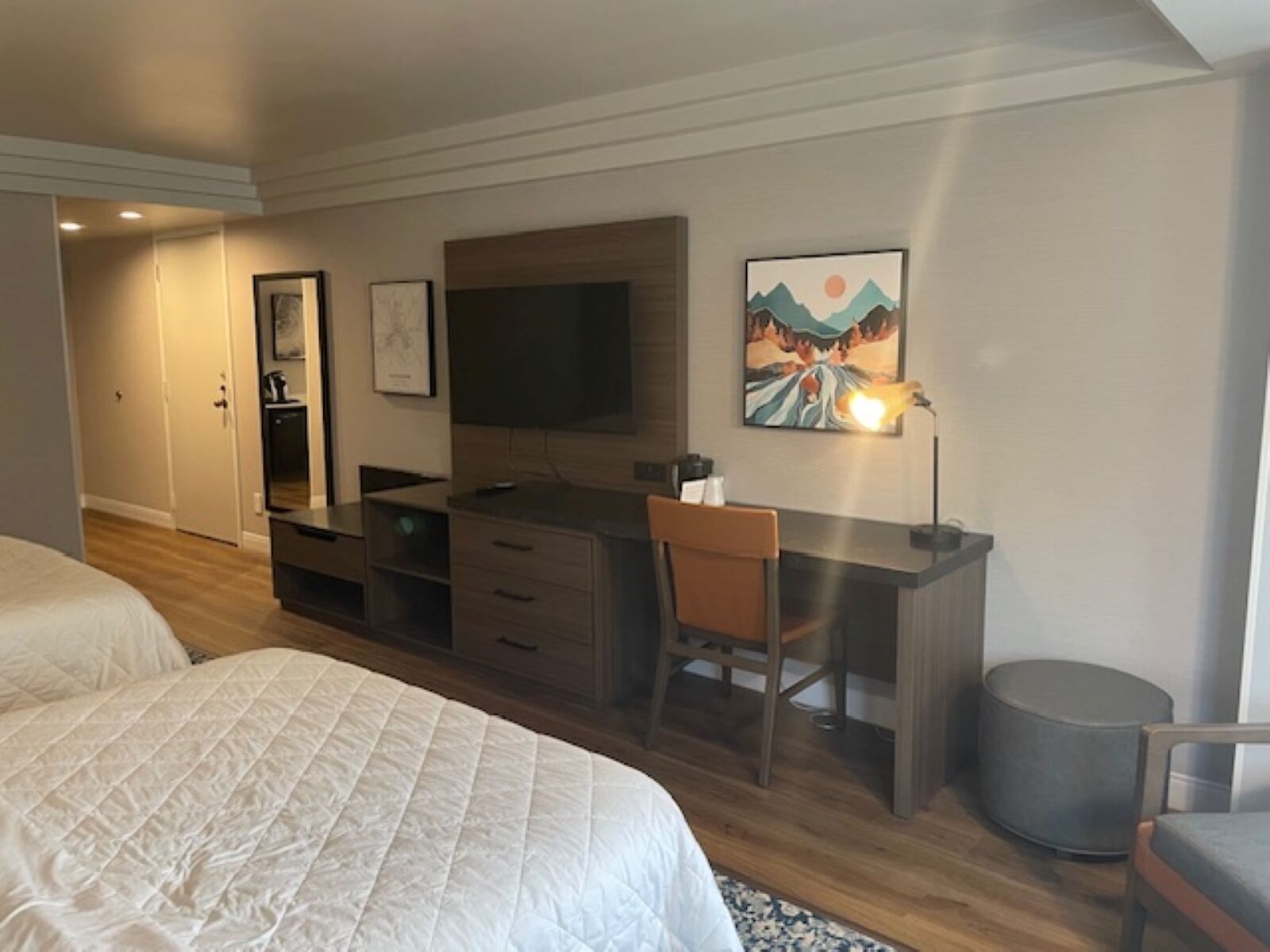 A hotel room with a bed, a desk with a chair, a TV, and artwork on the wall.