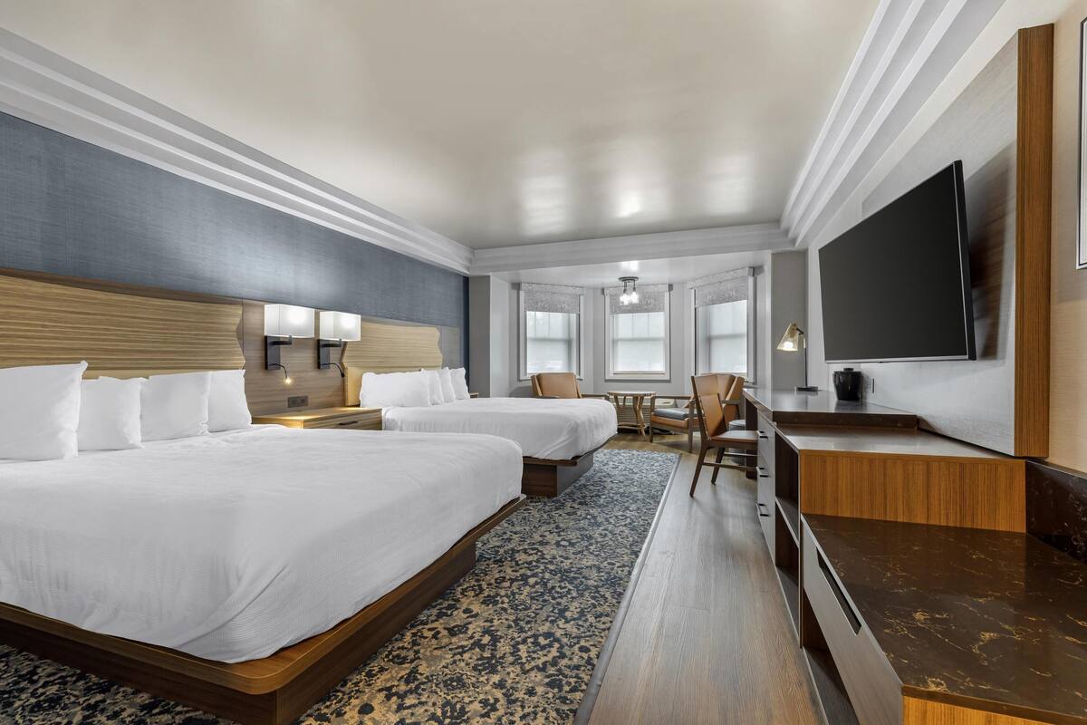 A hotel room with two beds, a desk, a flat-screen TV, and a window with a view.