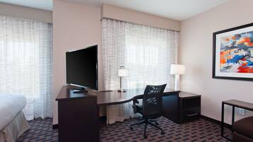A hotel room with a bed, a desk with a computer, a chair, a TV, a lamp, and a painting on the wall.
