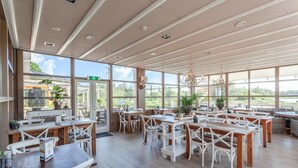 Un vaste espace extérieur aménagé pour les repas, avec des tables en bois et des chaises blanches, de grandes fenêtres offrant une vue sur la verdure et un plafond doté d’un éclairage encastré.