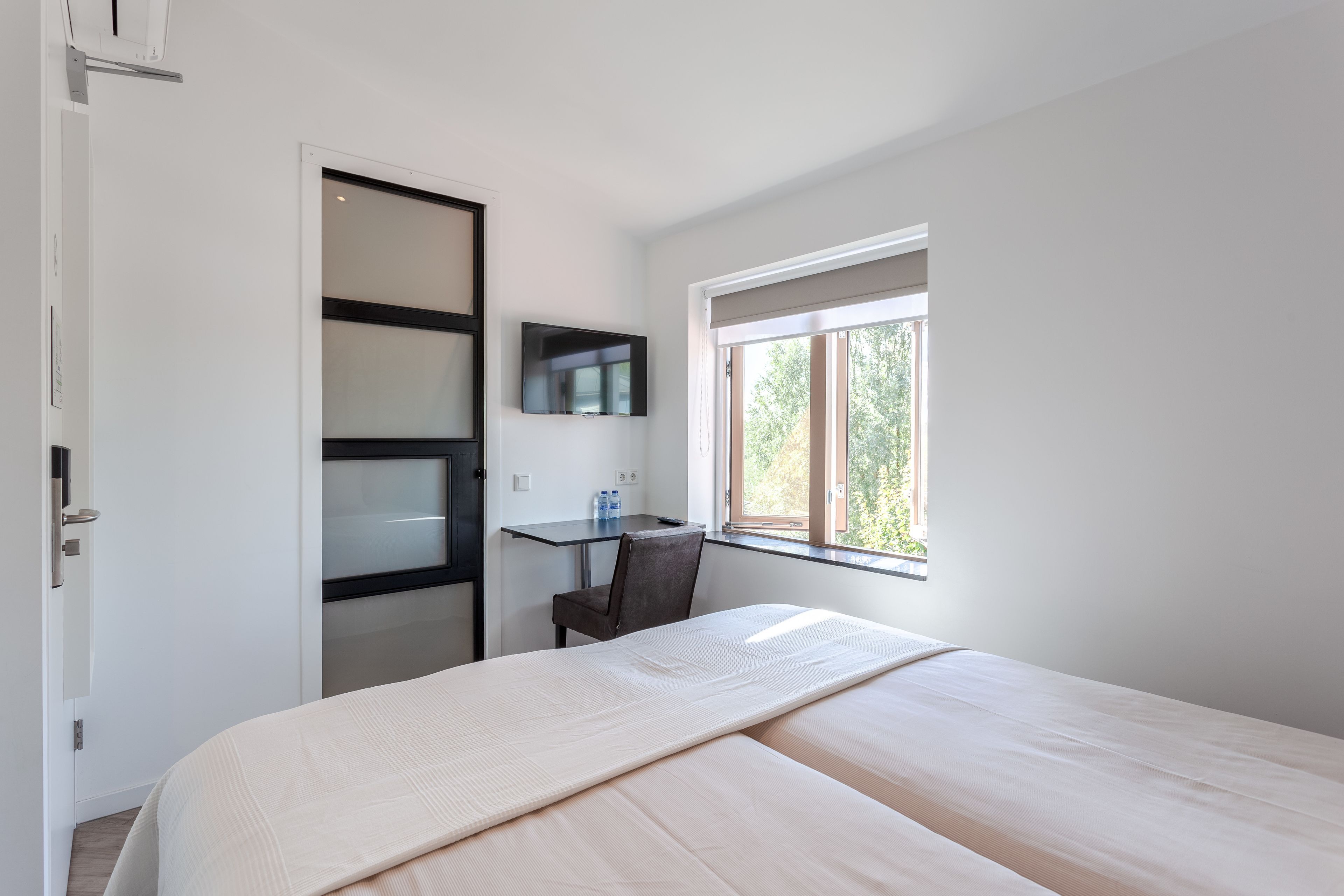 A bedroom with a bed, a desk, and a chair. There is a window providing natural light.