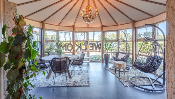 Un patio couvert avec un haut plafond, un lustre, un coin repas avec des chaises et des fauteuils suspendus.