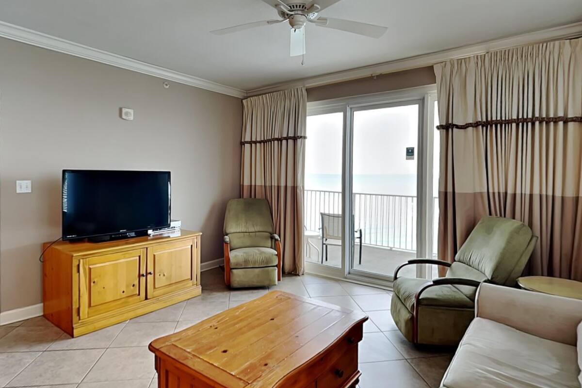 Uma sala de estar com televisão sobre um armário de madeira, duas poltronas, um sofá e um ventilador de teto.