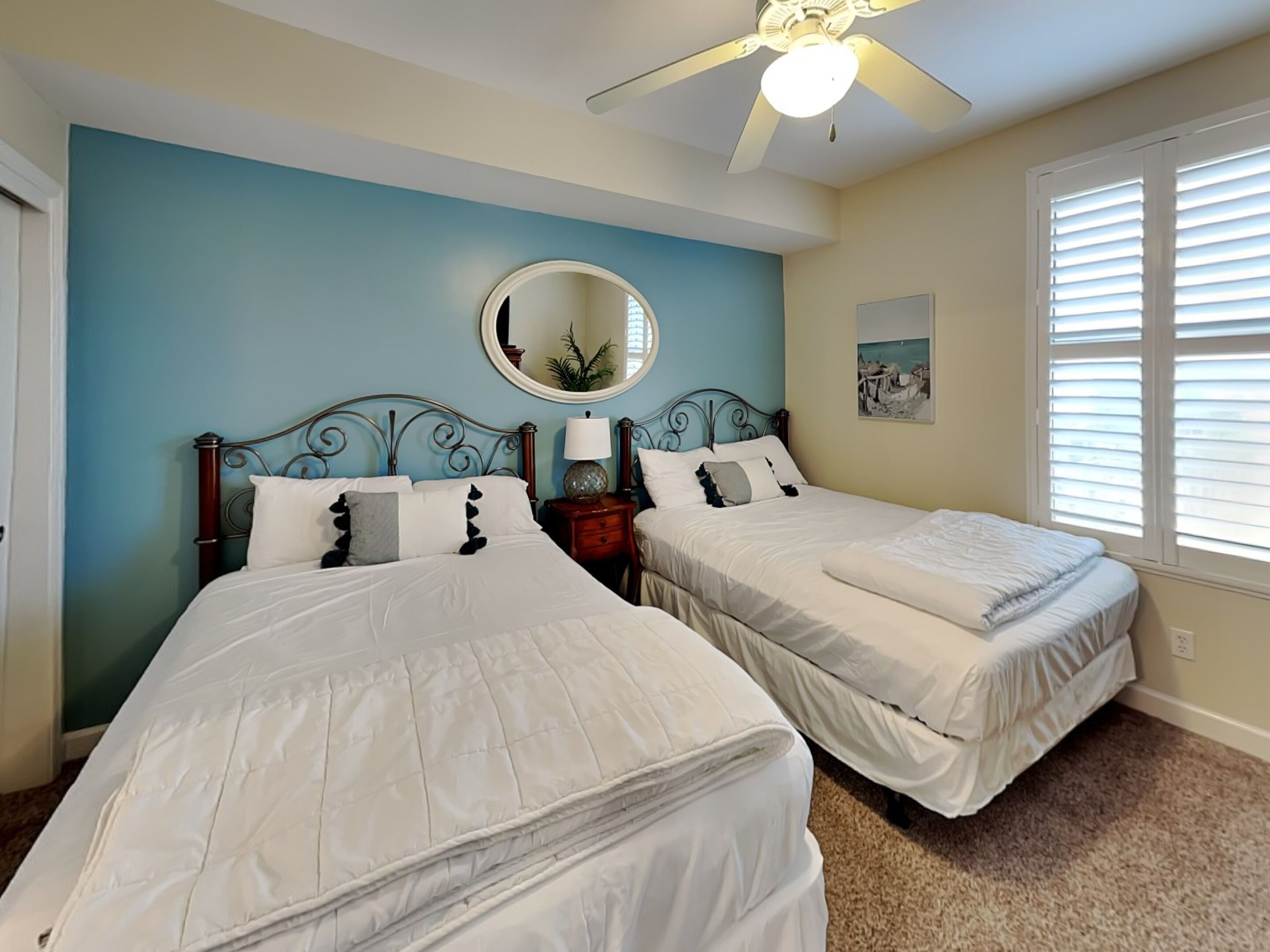 A bedroom with two beds, a ceiling fan, a round mirror, and a window with blinds.