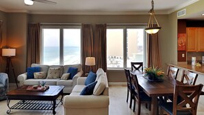 Ein Wohnzimmer mit einer Couch, einem Couchtisch, einem Esstisch und Stühlen. Große Fenster mit Vorhängen, ein Deckenventilator und eine Hängeleuchte sind ebenfalls vorhanden.