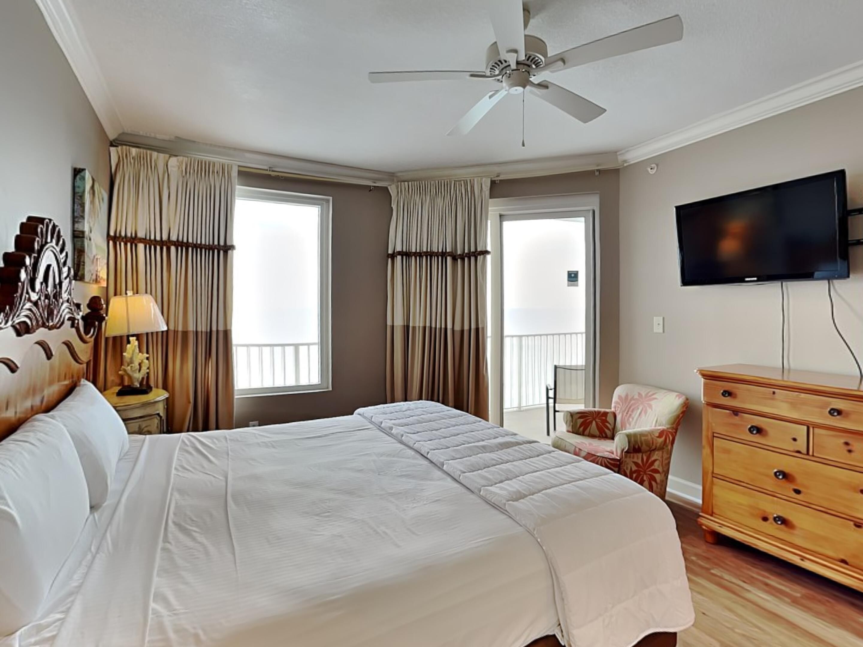 A hotel room with a large bed, a ceiling fan, a television, and a balcony with curtains.