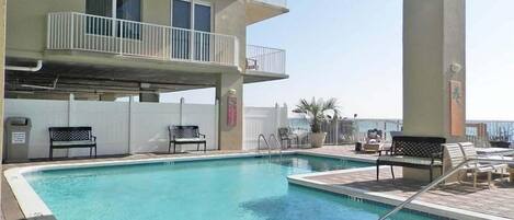 A swimming pool with a tiled deck, surrounded by lounge chairs and a building with balconies.