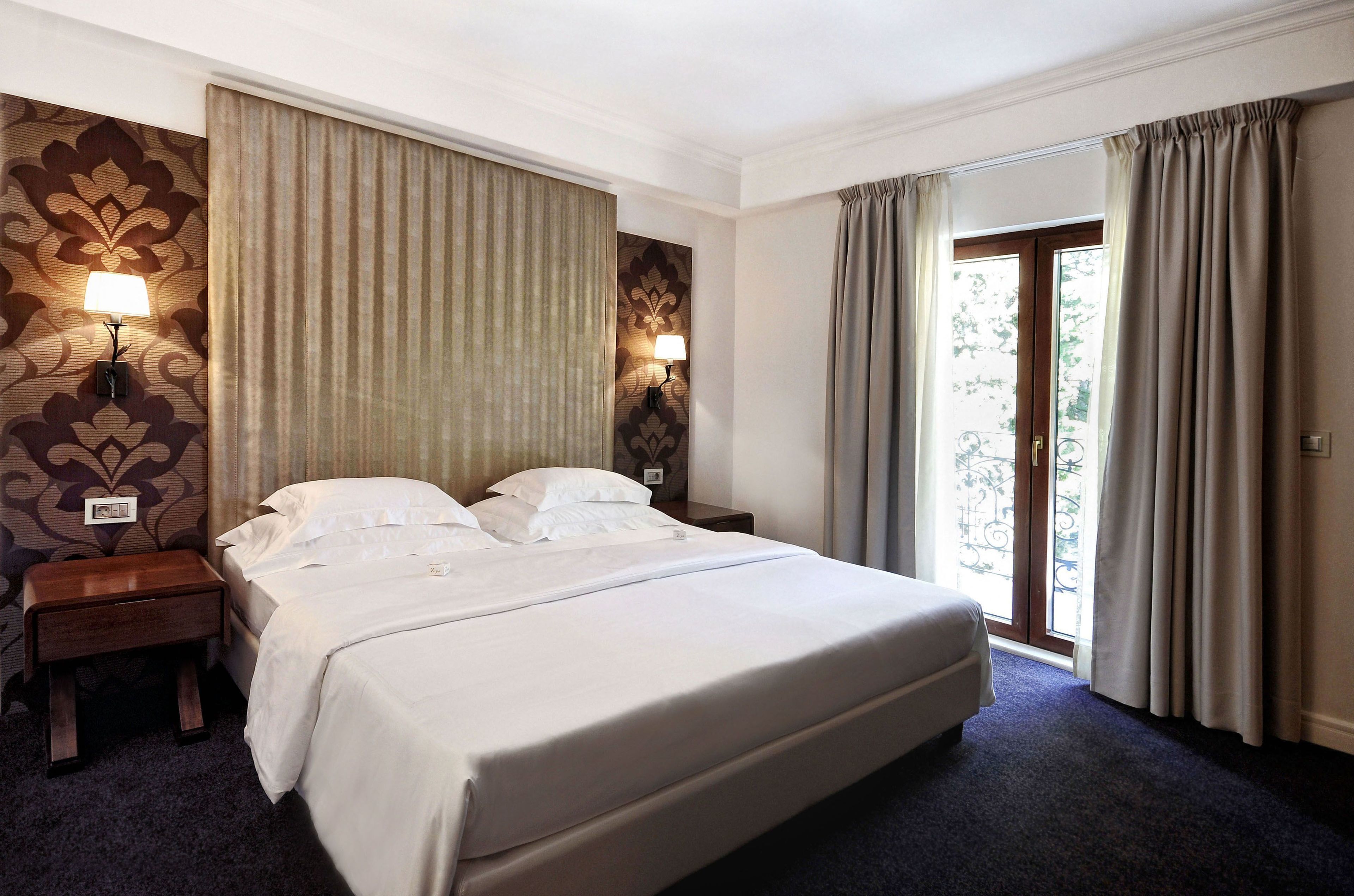A hotel room with a large bed, two bedside tables, a nightstand, and a window with curtains.