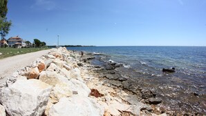 A rocky shoreline with a clear view of the sea, a paved path, and buildings in the background. - Villa Badi (Umag)