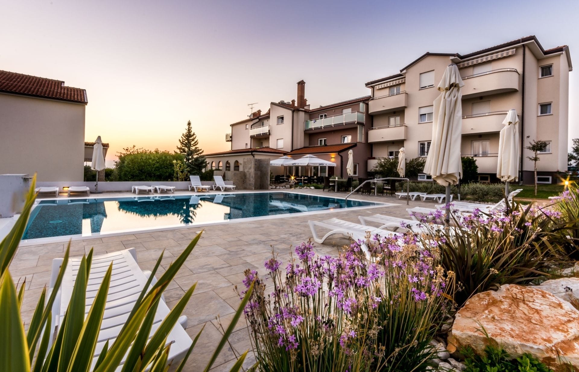 Una piscina con sillones de descanso, rodeada por un jardín con flores moradas y un edificio con balcones.