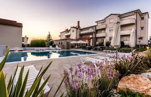 Una piscina con sillones de descanso, rodeada por un jardín con flores moradas y un edificio con balcones.