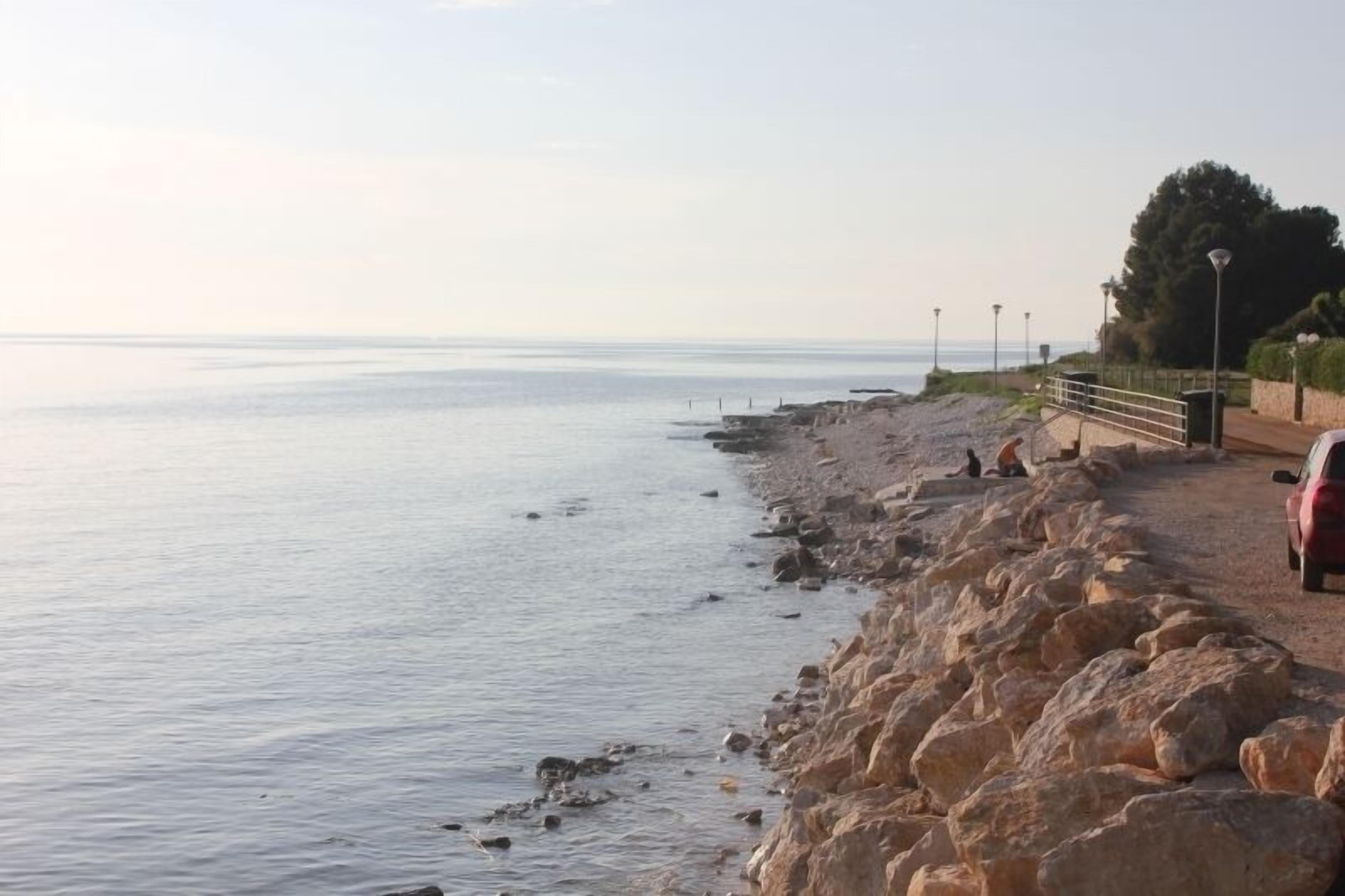 A coastal road with a rocky embankment, a parked car, and street lamps.