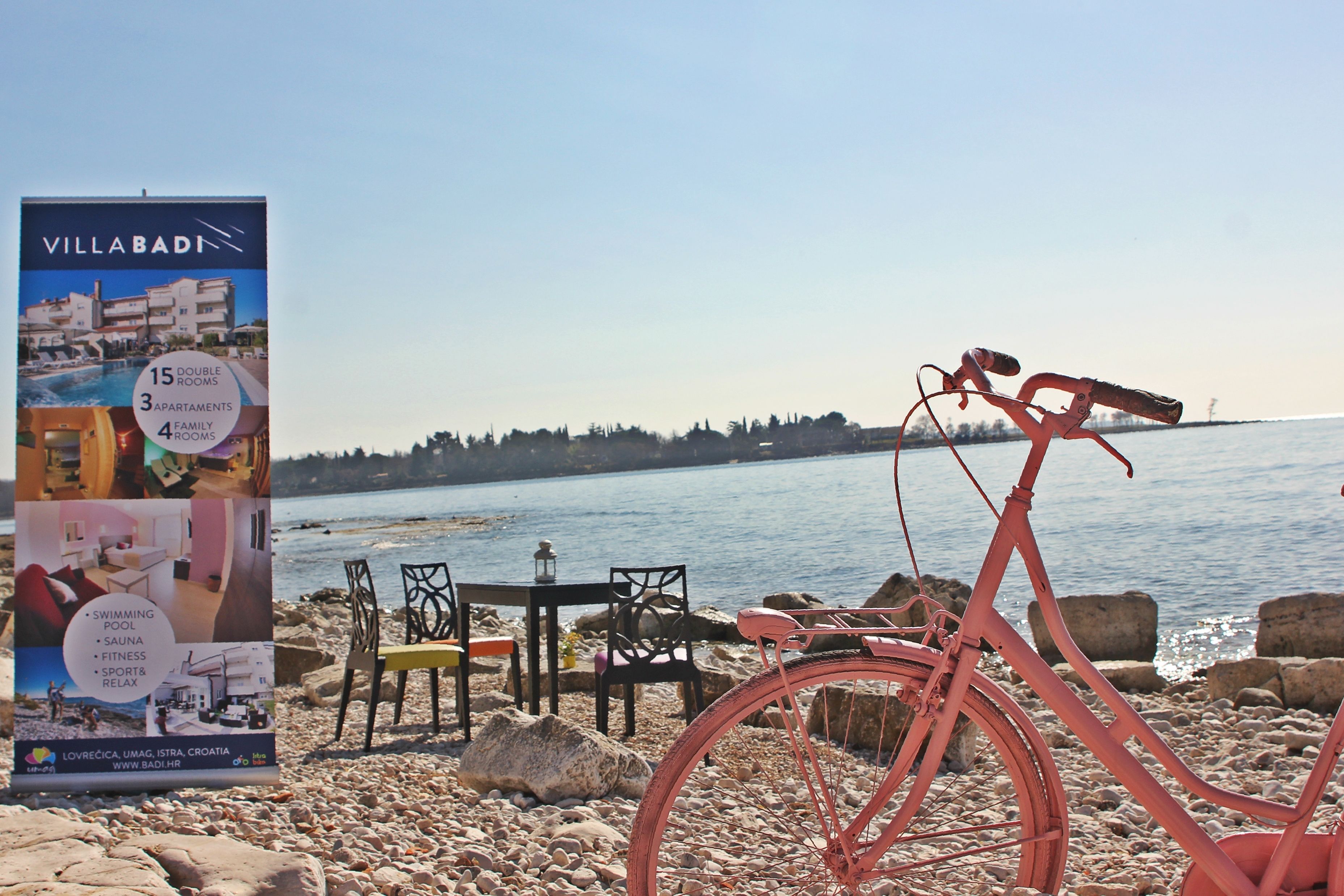 Una bicicleta roja estacionada cerca de una playa, con un cartel publicitario de Villa Badi, que ofrece comodidades como piscina, sauna, gimnasio y habitaciones familiares.