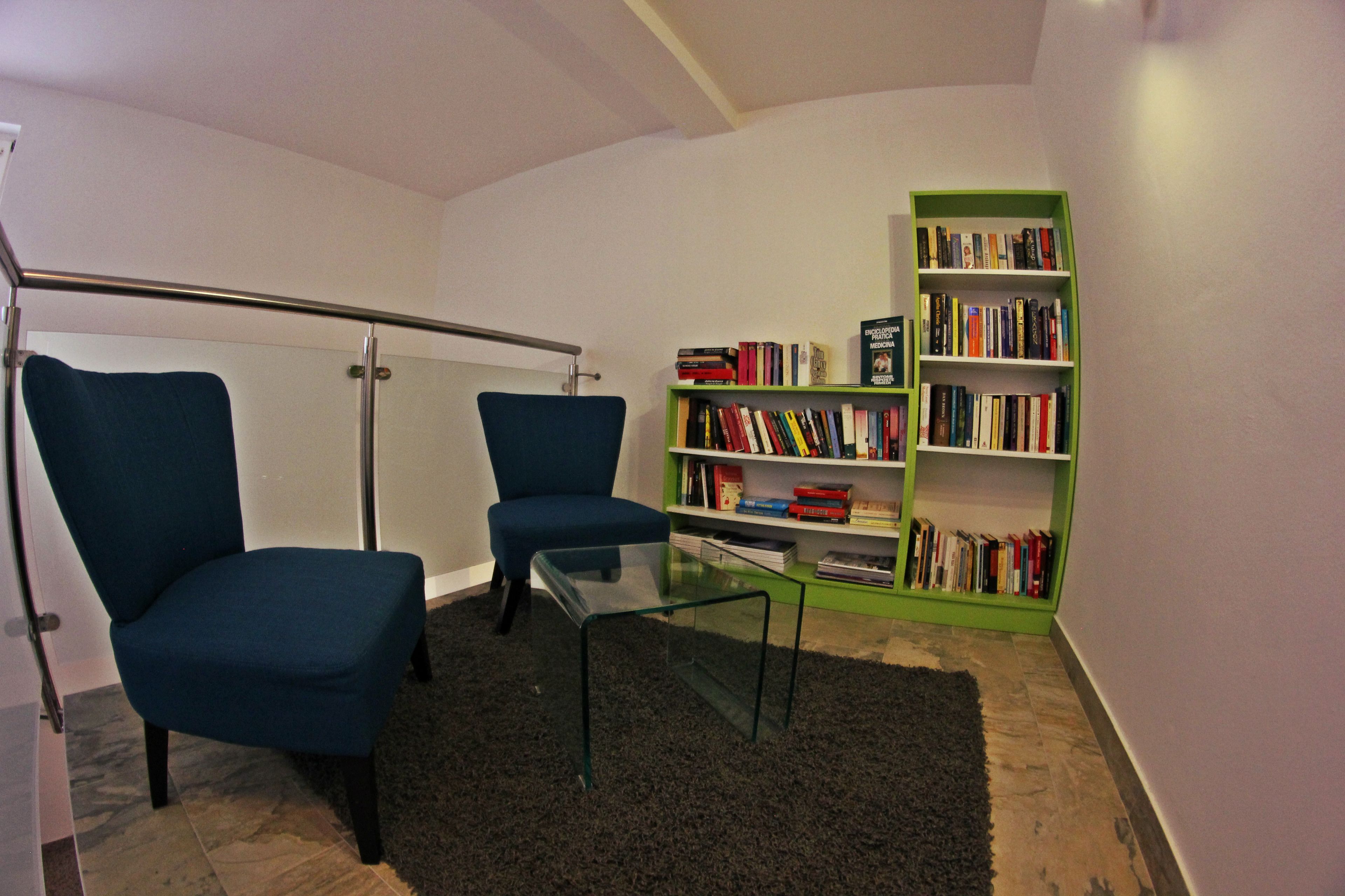 A modern living room with two blue armchairs, a glass coffee table, a green bookshelf, and a glass door leading to a balcony.