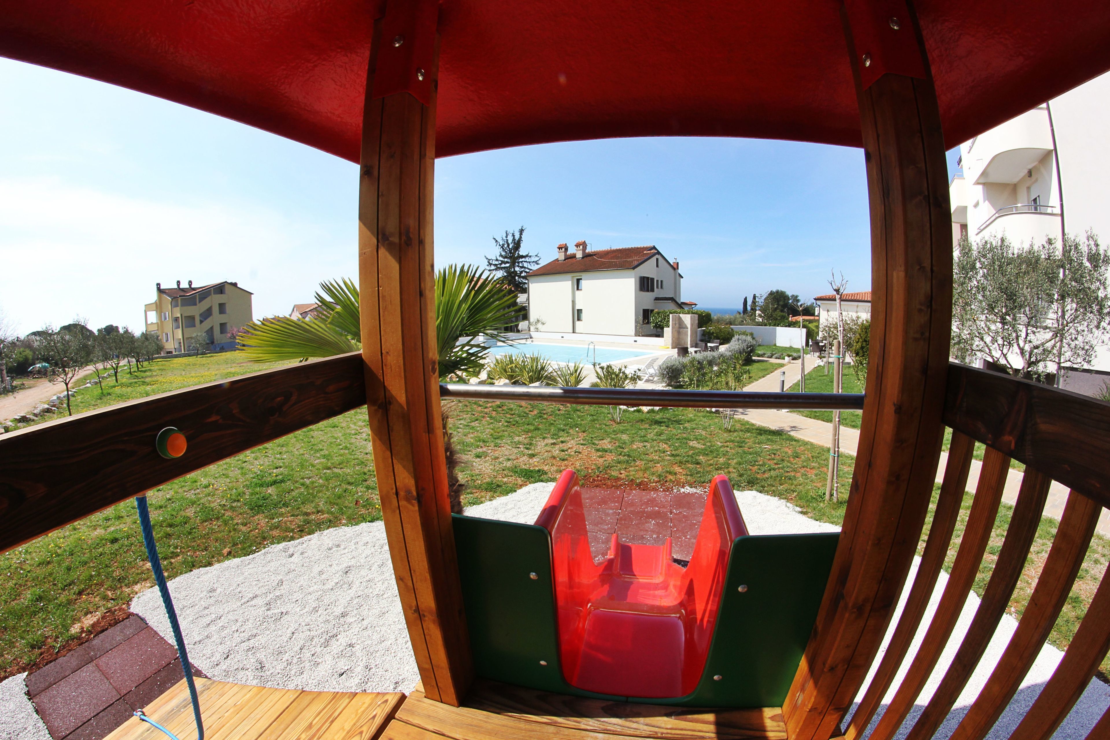A wooden deck with a red slide, overlooking a swimming pool and a white building with a red roof.