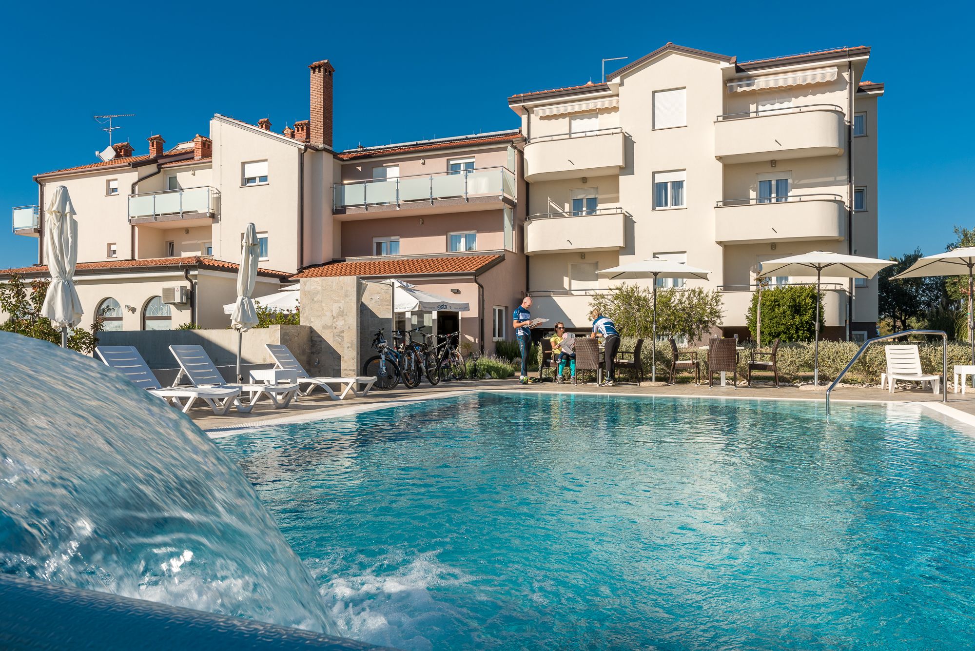 Una piscina con sillones de descanso y sombrillas, rodeada de edificios de varios pisos con balcones.