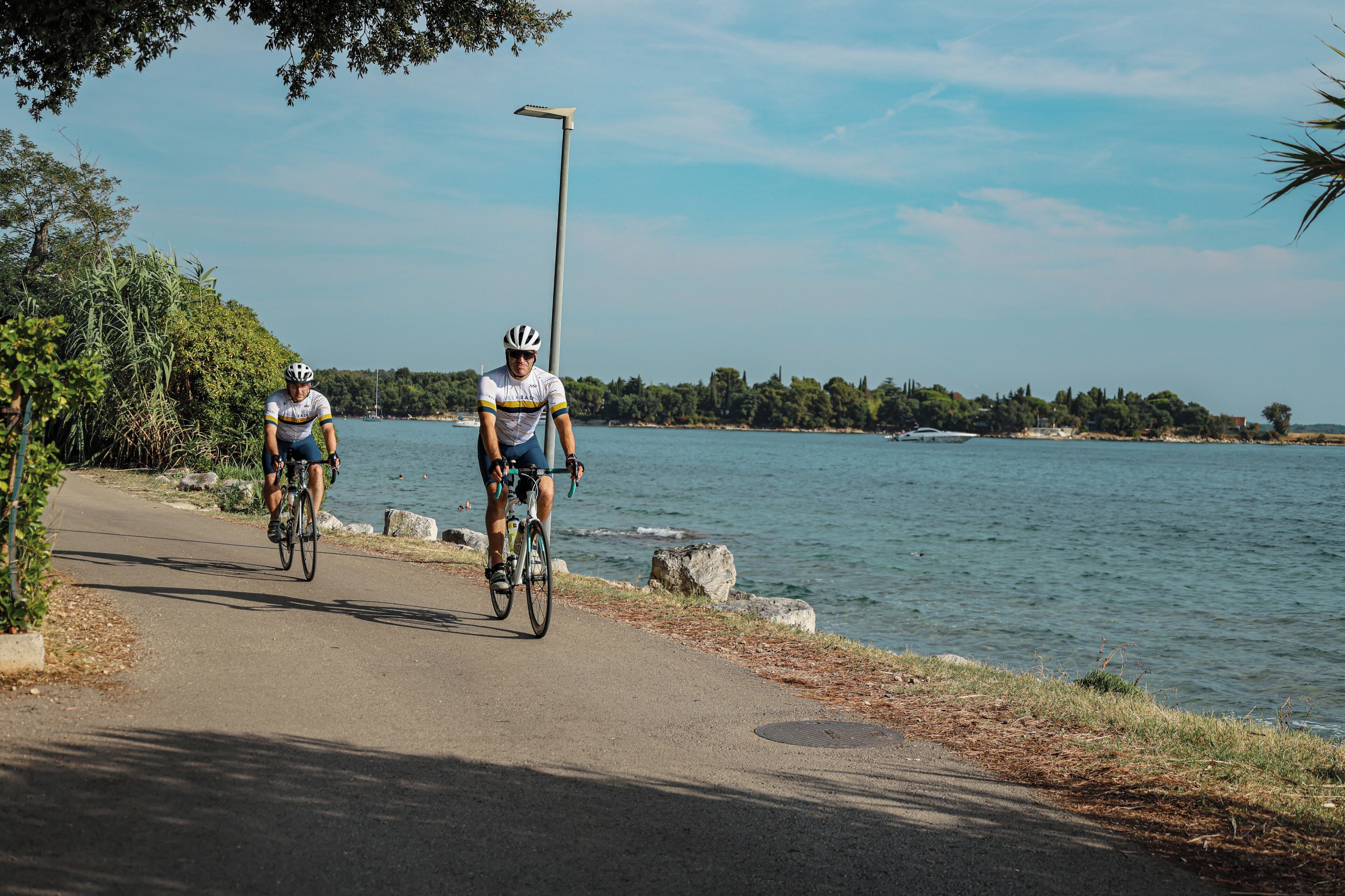 Dois ciclistas em um caminho pavimentado ao lado de um corpo d'água, com árvores e céu claro ao fundo.
