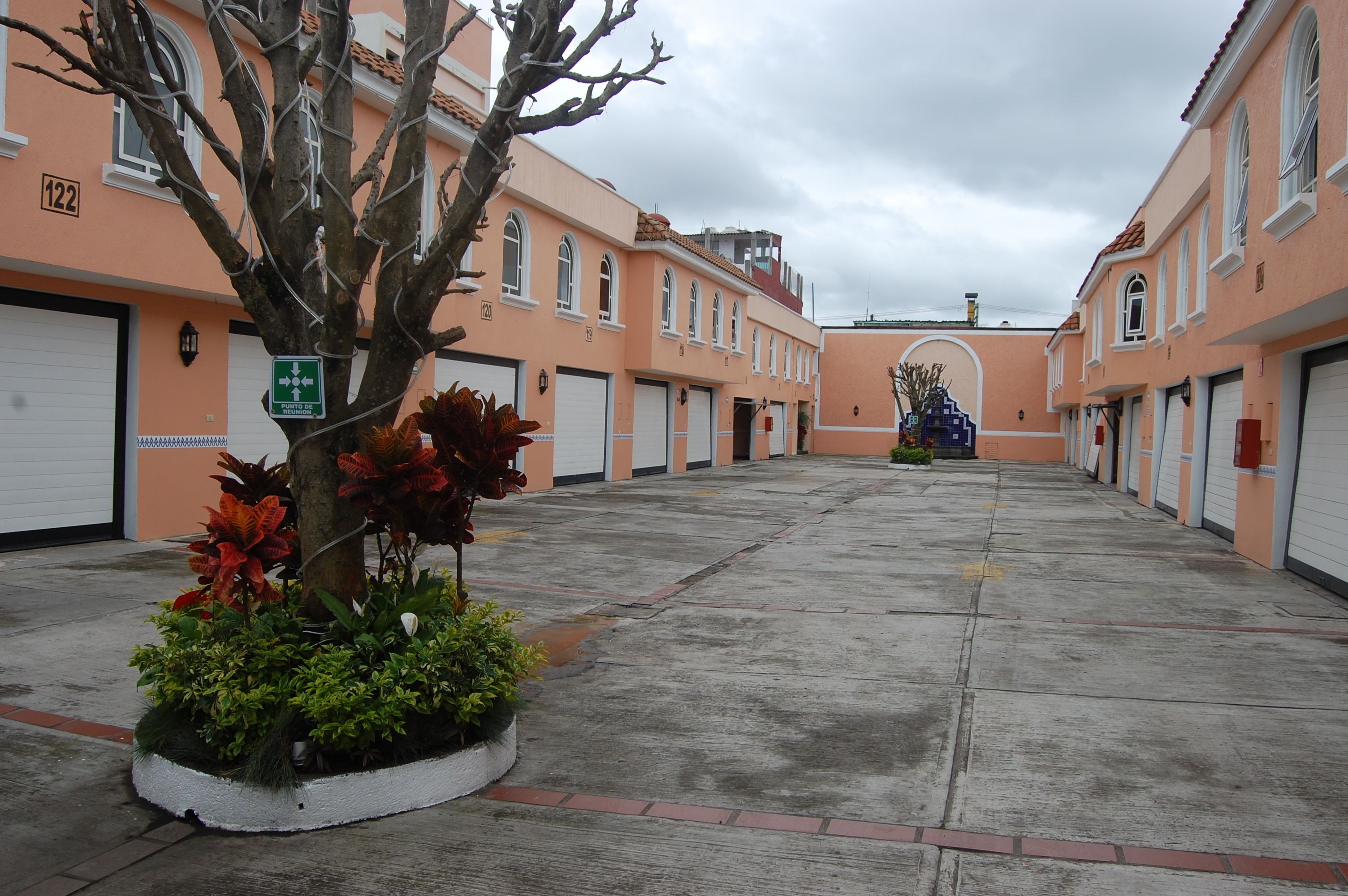 A building with a row of garage doors, a tree with red flowers, and a sign indicating a parking area.