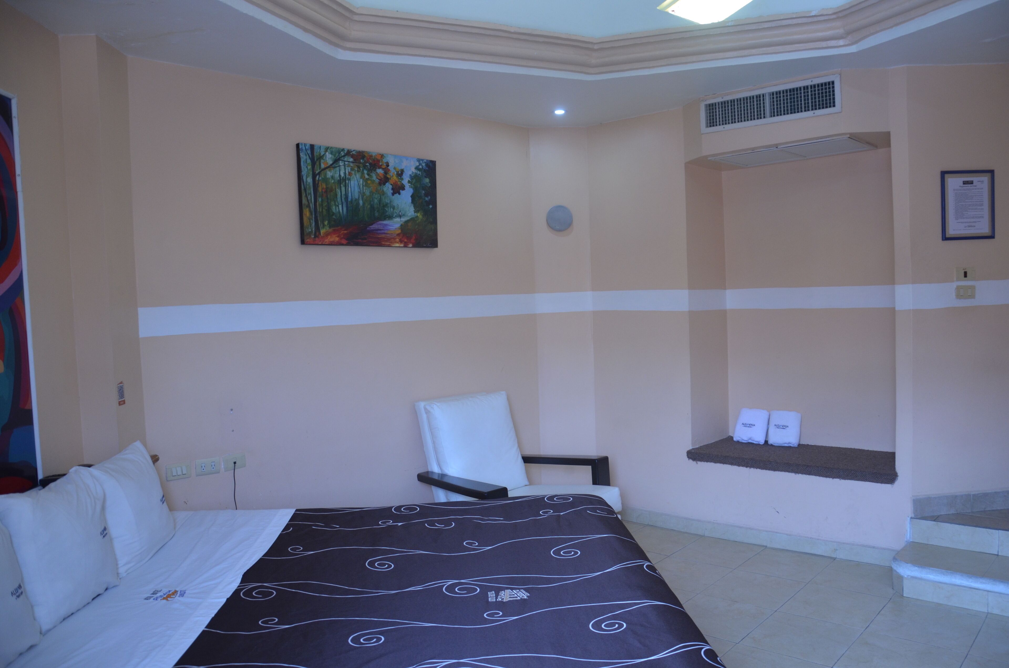 Una habitación de hotel con una cama, una silla, un cuadro en la pared, un estante con toallas y una escalera.