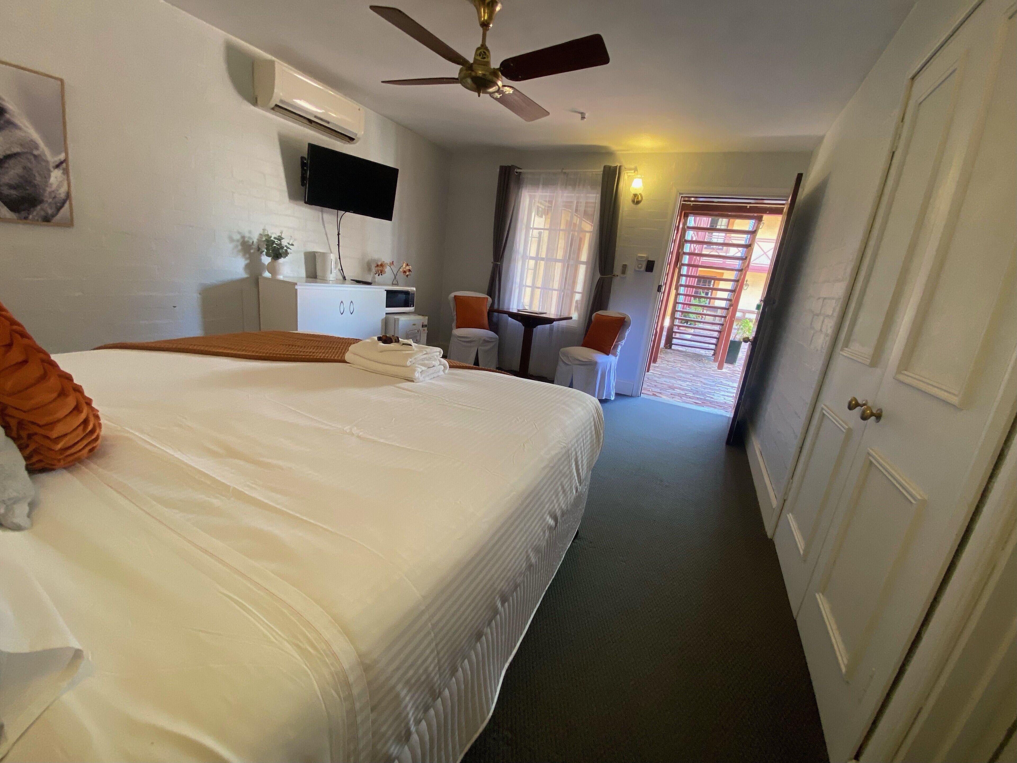 A hotel room with a large bed, a ceiling fan, a flat-screen TV, a desk, and a small table. There is a door leading to a balcony.