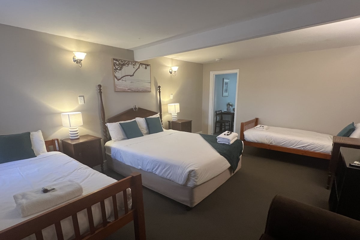 A hotel room with two single beds, a double bed, a wooden chair, a desk, and a nightstand with a lamp.