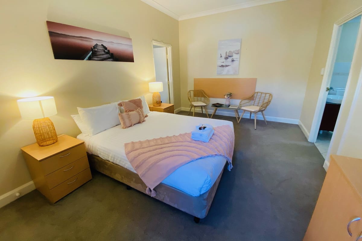 A hotel room with a bed, bedside tables, a desk, two chairs, and artwork on the walls.
