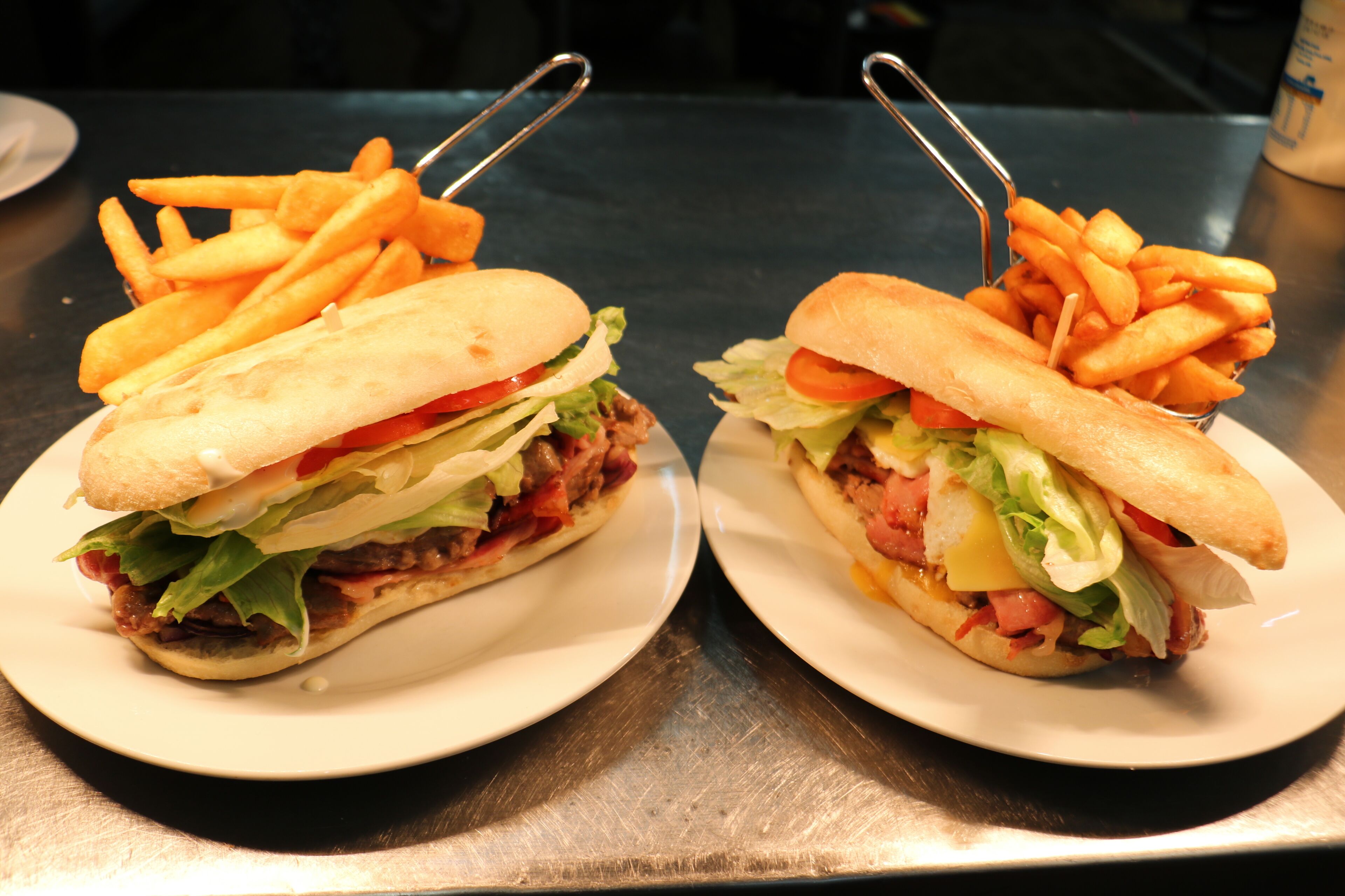 To sandwicher med pommes frites på hvide tallerkener på et metalbord.