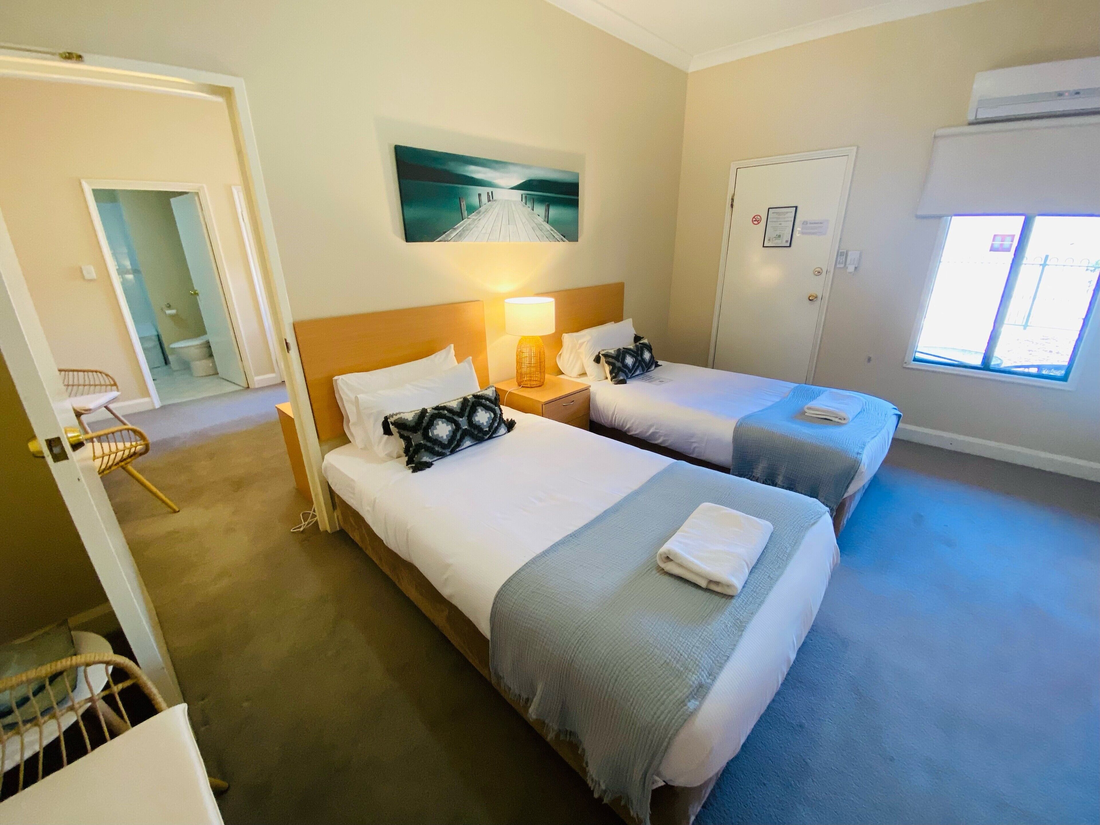 A hotel room with two beds, a painting above the headboard, a window with blinds, and a door with a sign.
