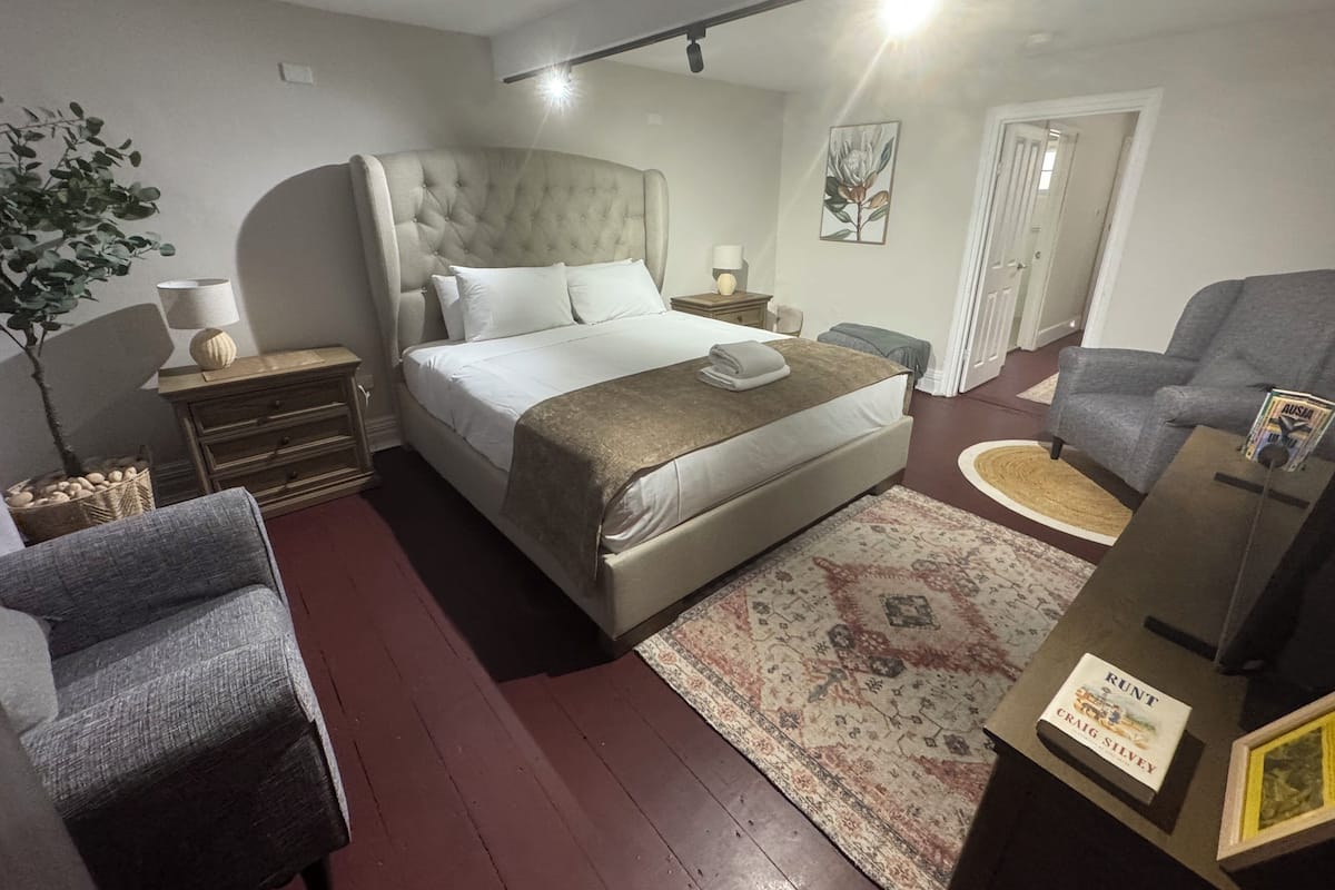 A bedroom with a large bed, a bedside table with a lamp, a chair, a small table with a book, and a door.