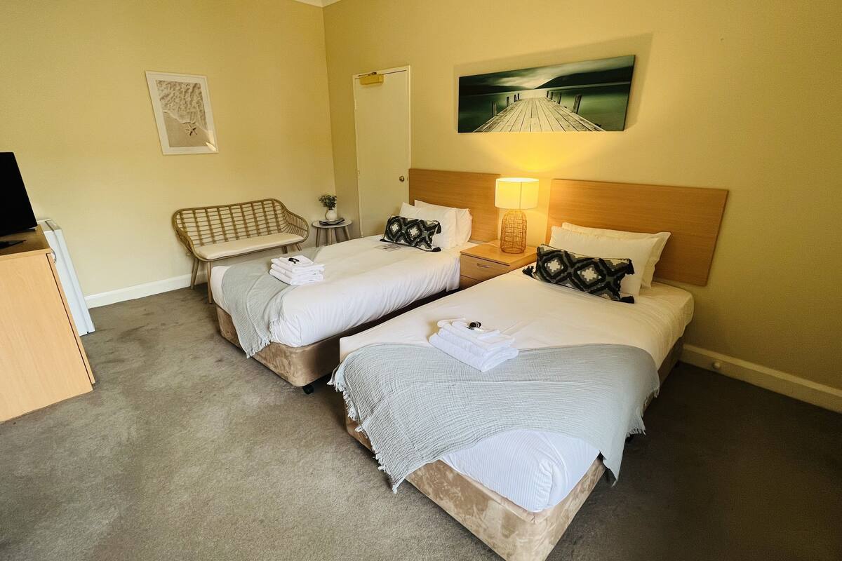 A hotel room with two beds, a TV, a bench, a small table, and a painting on the wall.