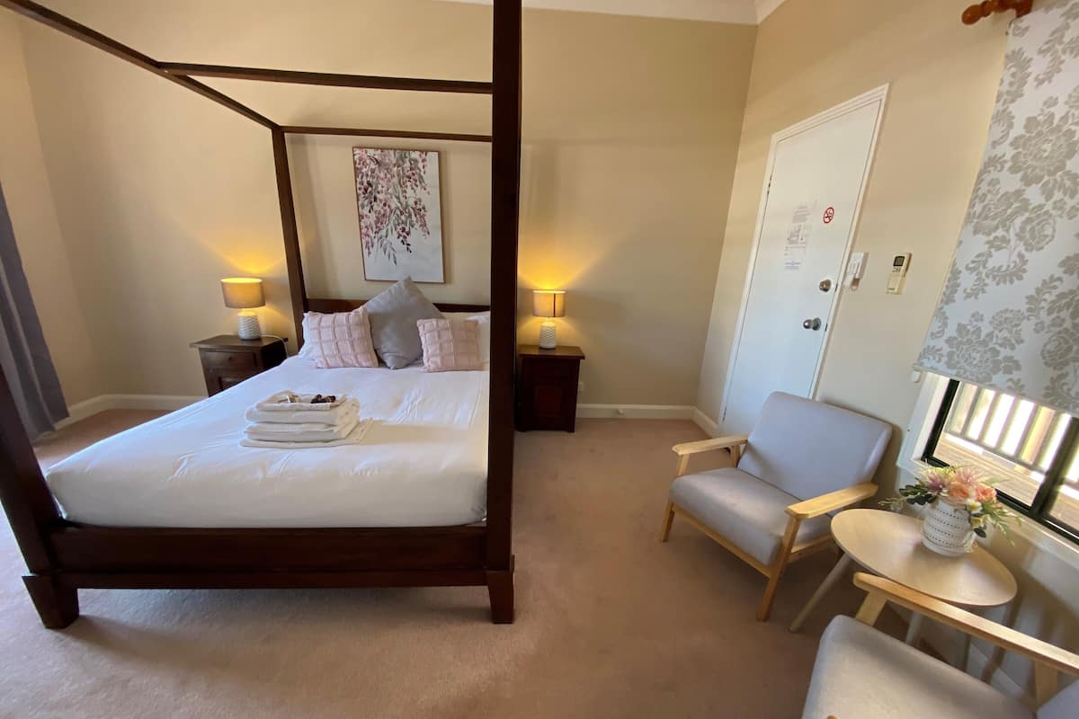 A four-poster bed with a headboard, two bedside tables with lamps, a chair, a small table, and a window with a view.