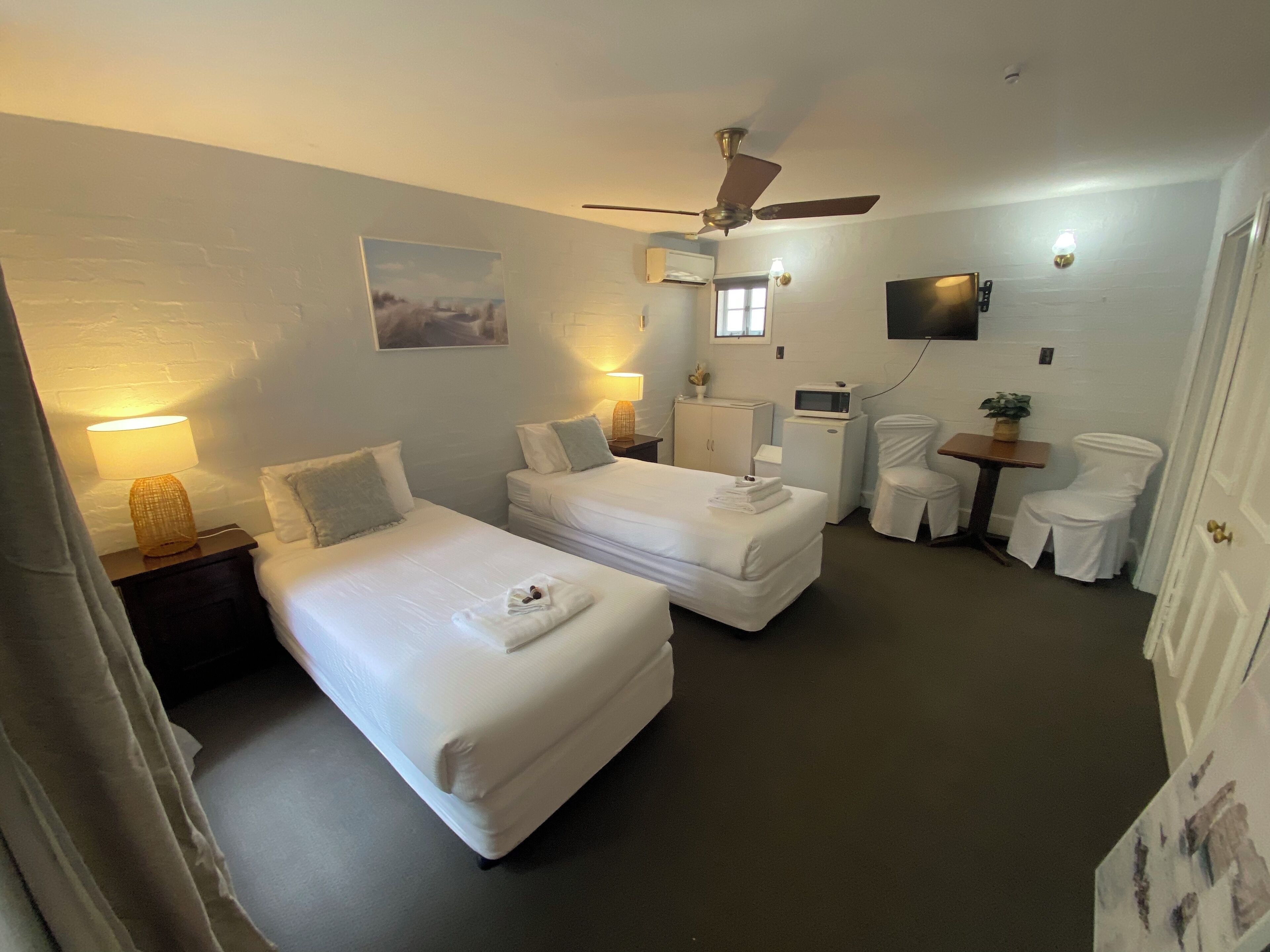 A hotel room with two single beds, a ceiling fan, a flat-screen TV, a microwave, a small table with a plant, and a white chair.