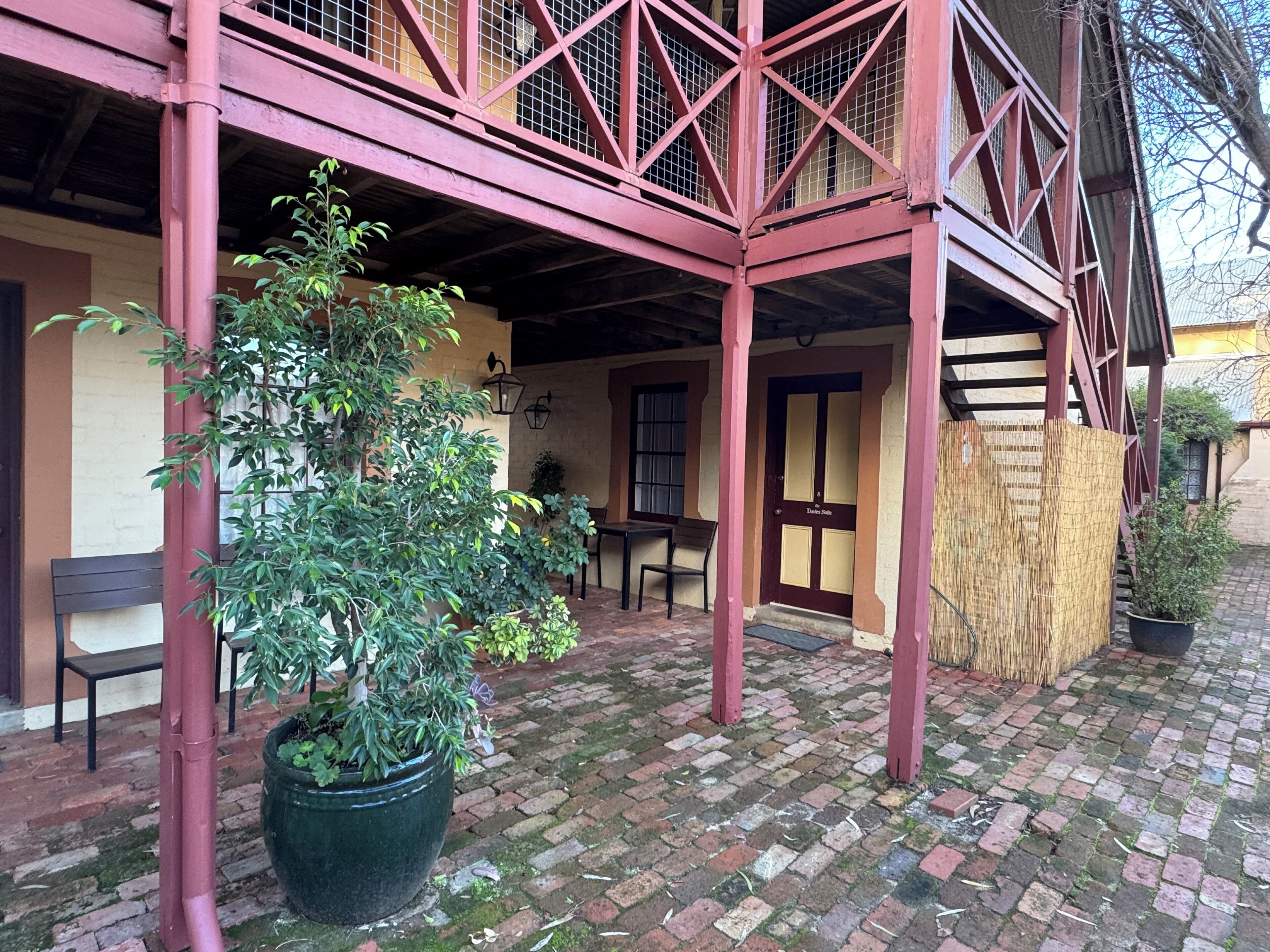 A covered patio area with a brick floor, a large potted plant, a wooden bench, and a table.
