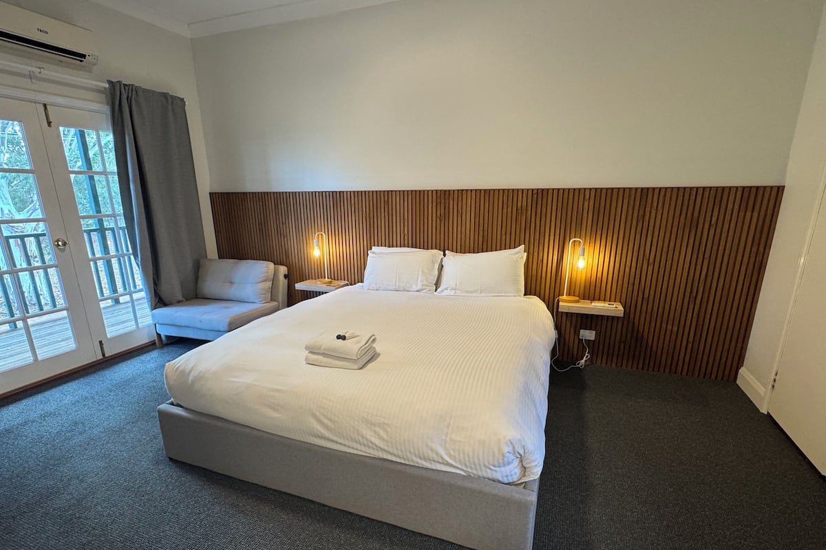 A hotel room with a large bed, a chair, a desk with a lamp, and a view of the outdoors through a door.