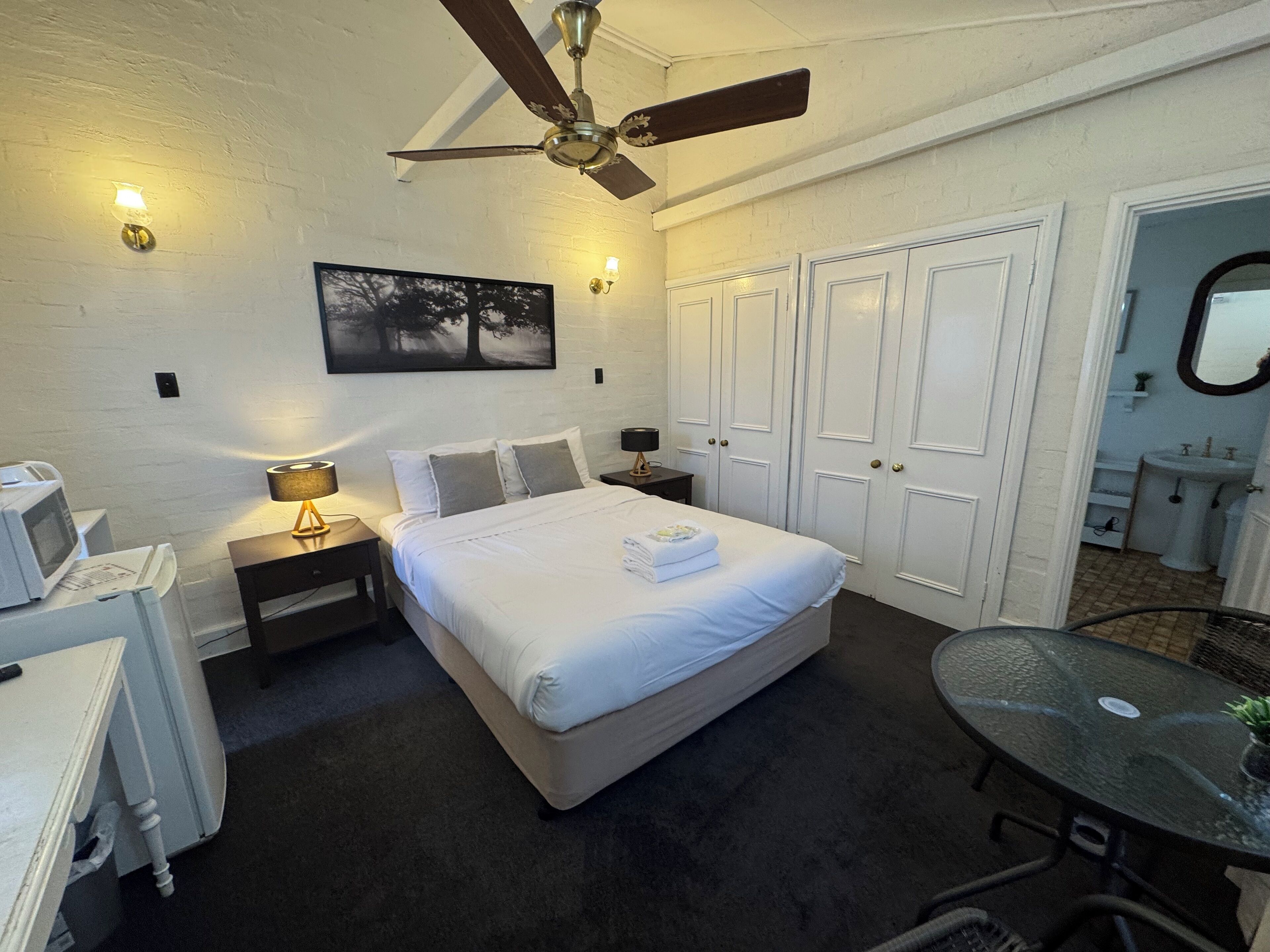 A bedroom with a bed, bedside tables, a ceiling fan, a painting, and a bathroom visible through an open door.