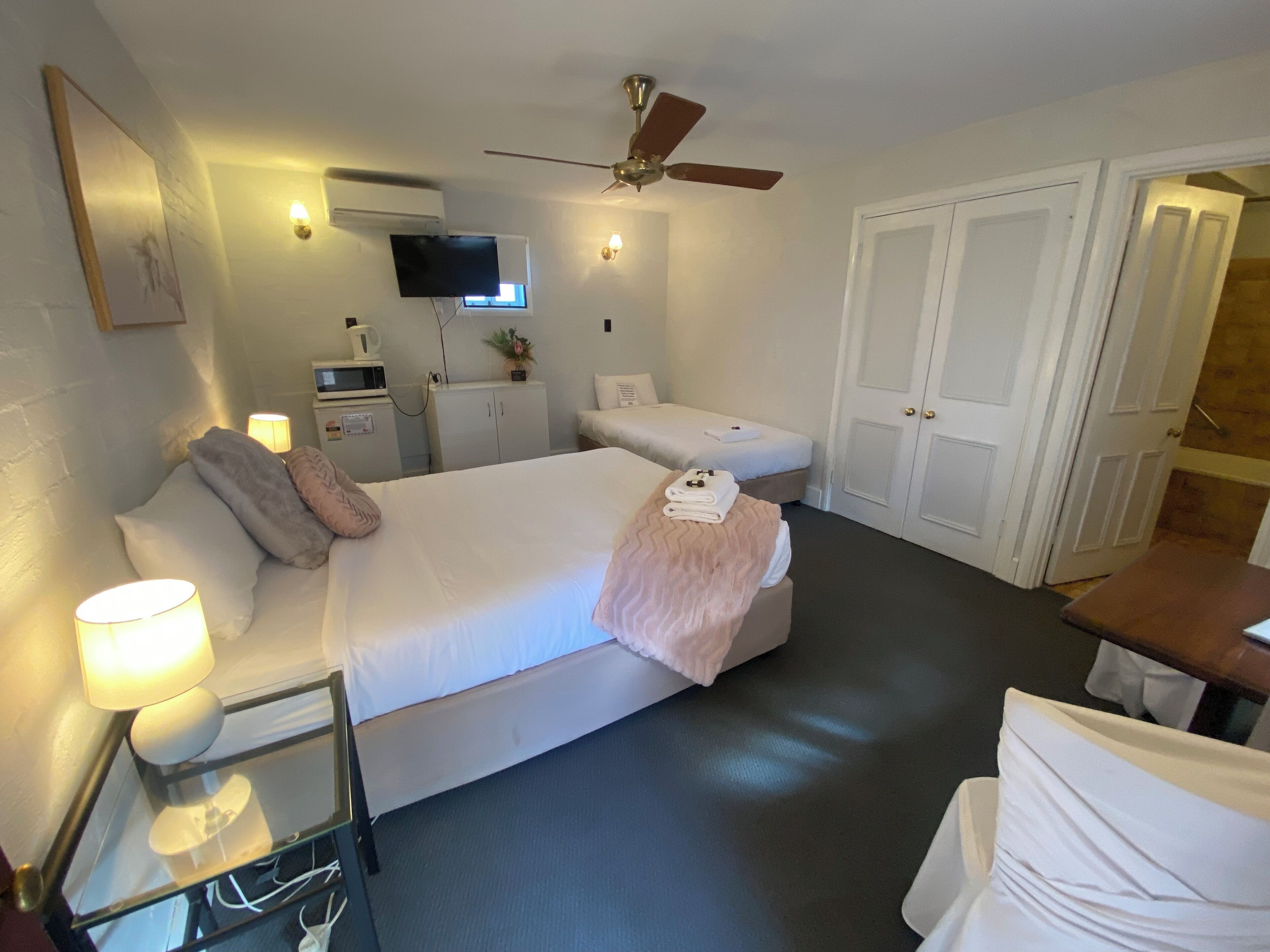 Habitación de hotel con cama, lámparas de noche, ventilador de techo, televisor, microondas, un mueble pequeño y una puerta.