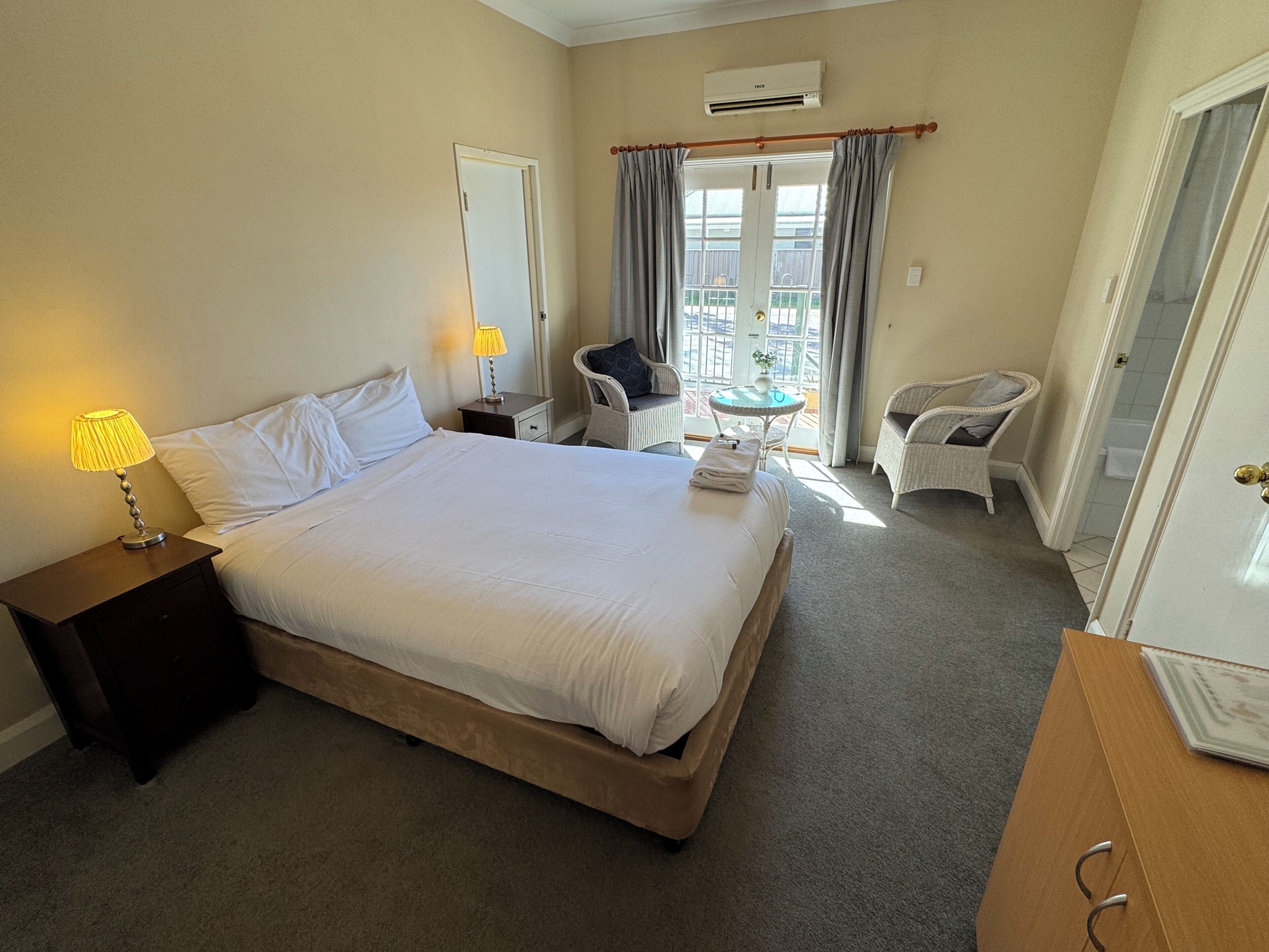 Habitación de hotel con una cama grande, una mesita de noche con lámpara, una silla, una pequeña mesa con un florero y un balcón con cortinas.