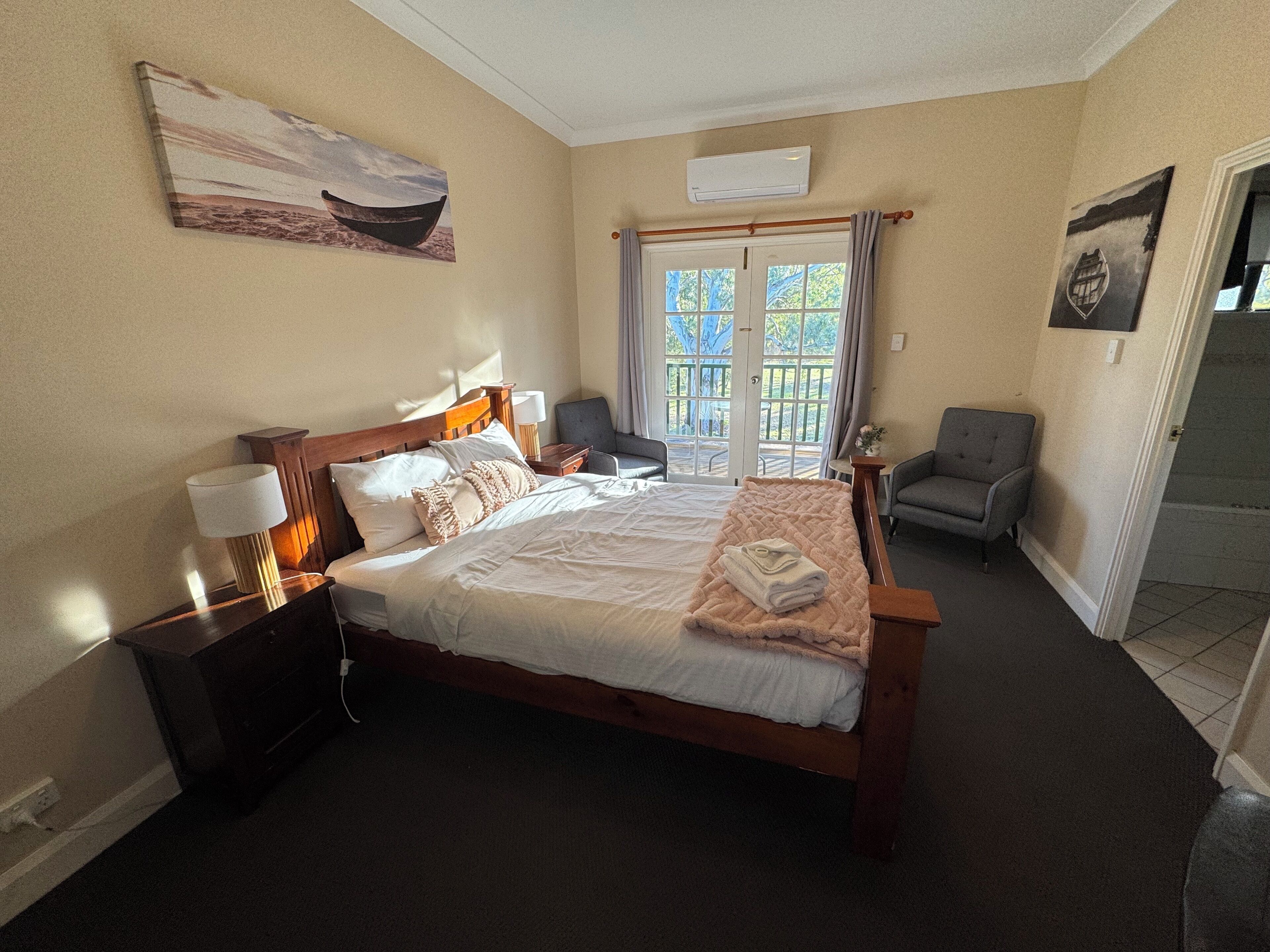 A neatly made bed with a wooden headboard, a bedside table with a lamp, and a chair in a room with a balcony view.