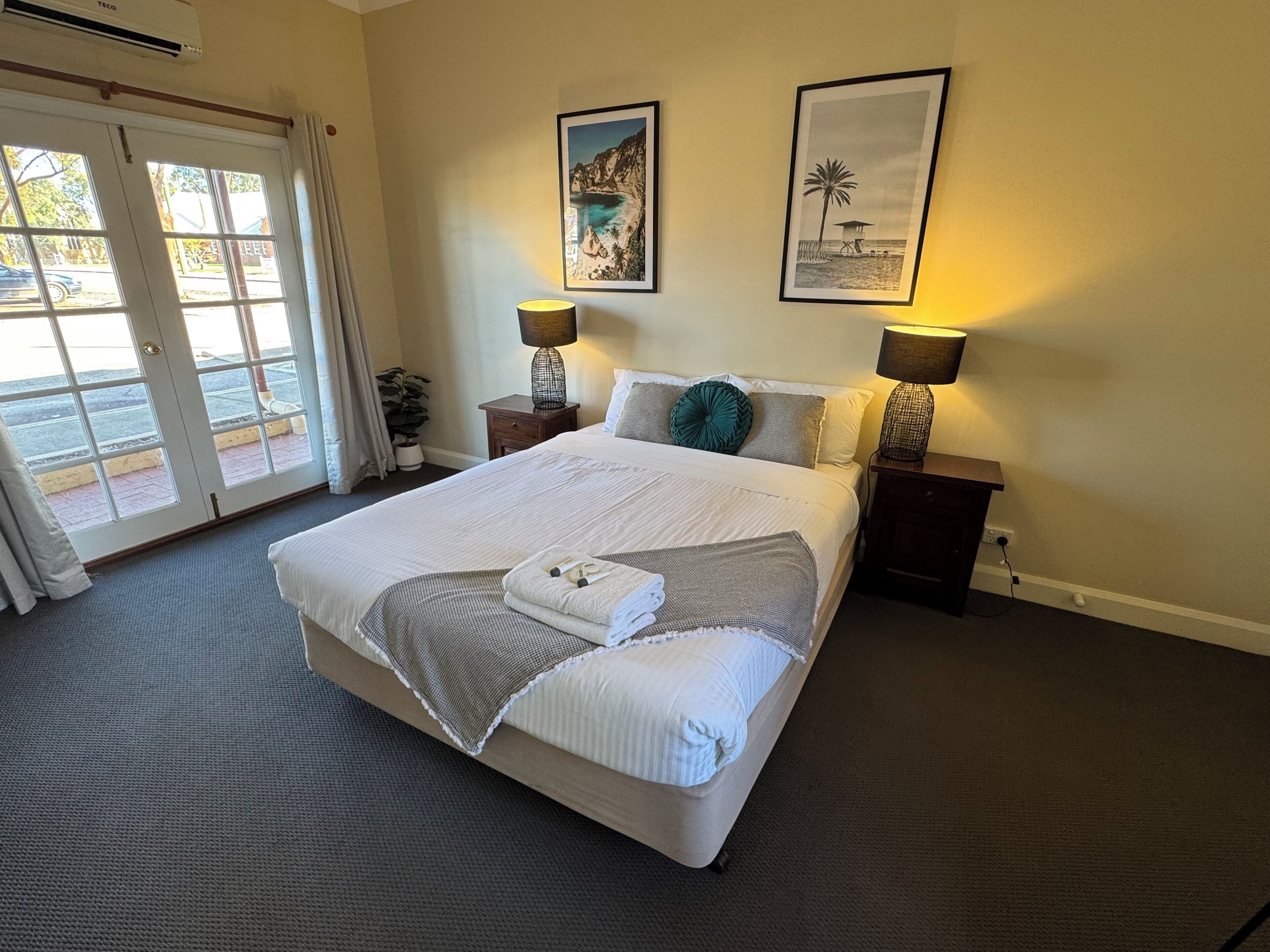 A hotel room with a large bed, two bedside lamps, a window with curtains, and two framed pictures on the wall.