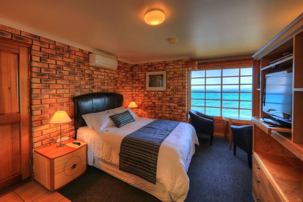 A bedroom with a brick wall, a bed with a headboard, bedside lamps, a desk with a chair, and a TV.