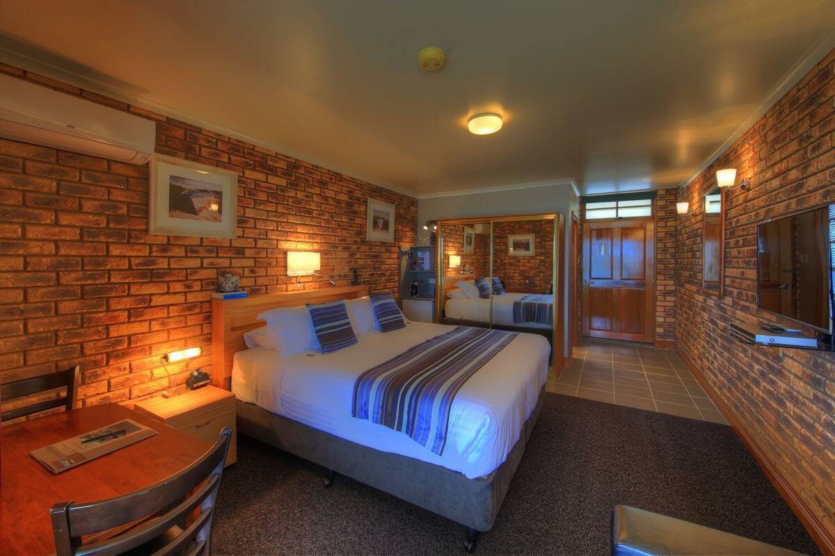 A hotel room with a brick wall, a bed with striped bedding, a desk with a chair, and a television.