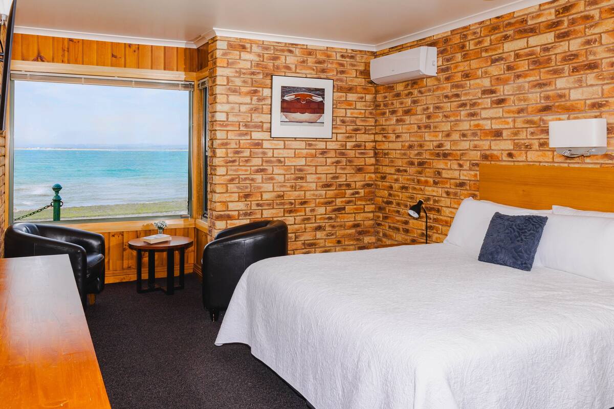 A hotel room with a brick wall, a large bed, a small table, a chair, and a view of the ocean.