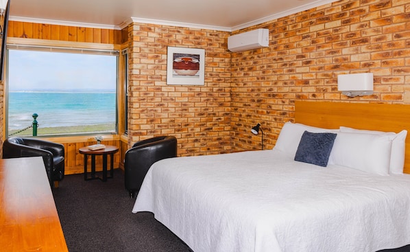Habitación de hotel con pared de ladrillo, una cama grande, una mesita, una silla y vista al océano.