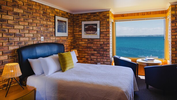 Habitación de hotel con pared de ladrillo, una cama grande con ropa blanca, cabecero negro y vista al mar a través de una ventana.