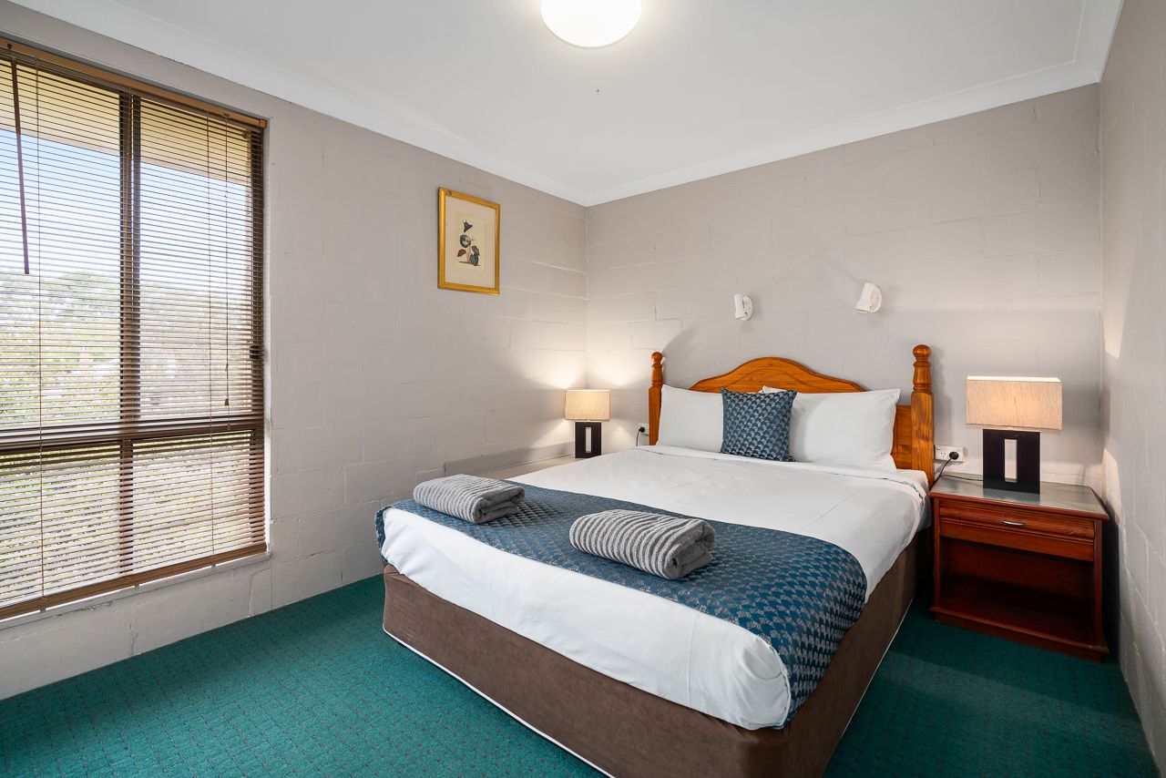 Une chambre d’hôtel avec un grand lit, deux tables de chevet, une fenêtre avec des stores, un tableau accroché au mur et une moquette verte.