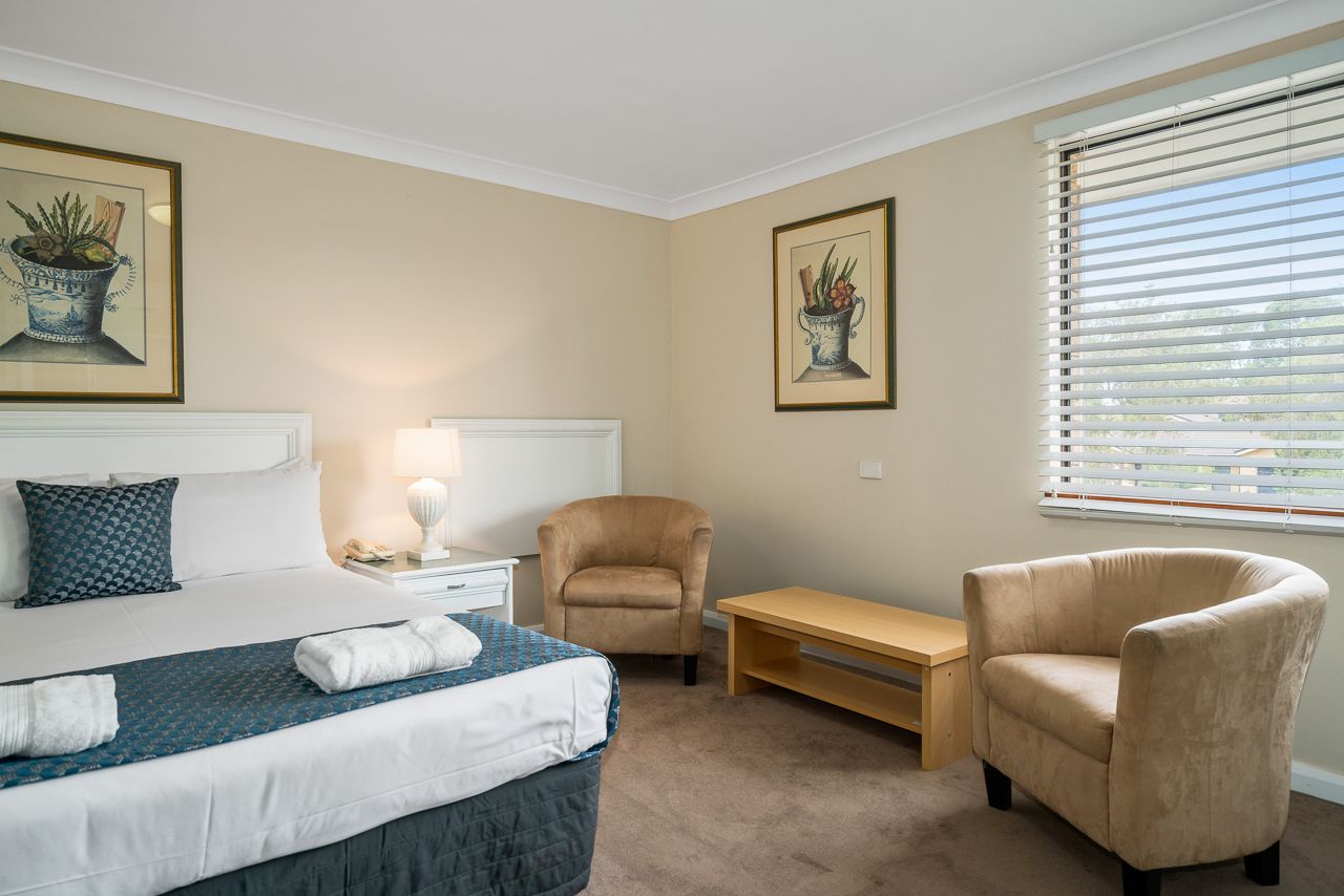 A hotel room with a bed, two armchairs, a nightstand with a lamp, and two framed pictures on the wall.