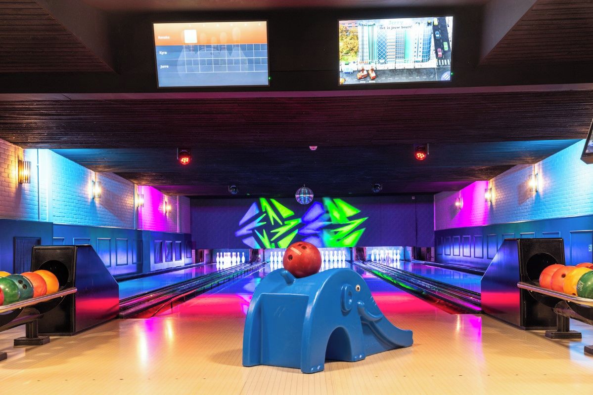 Bolera con múltiples pistas, una bola de bolos en un lanzador azul y una pantalla grande mostrando un juego.