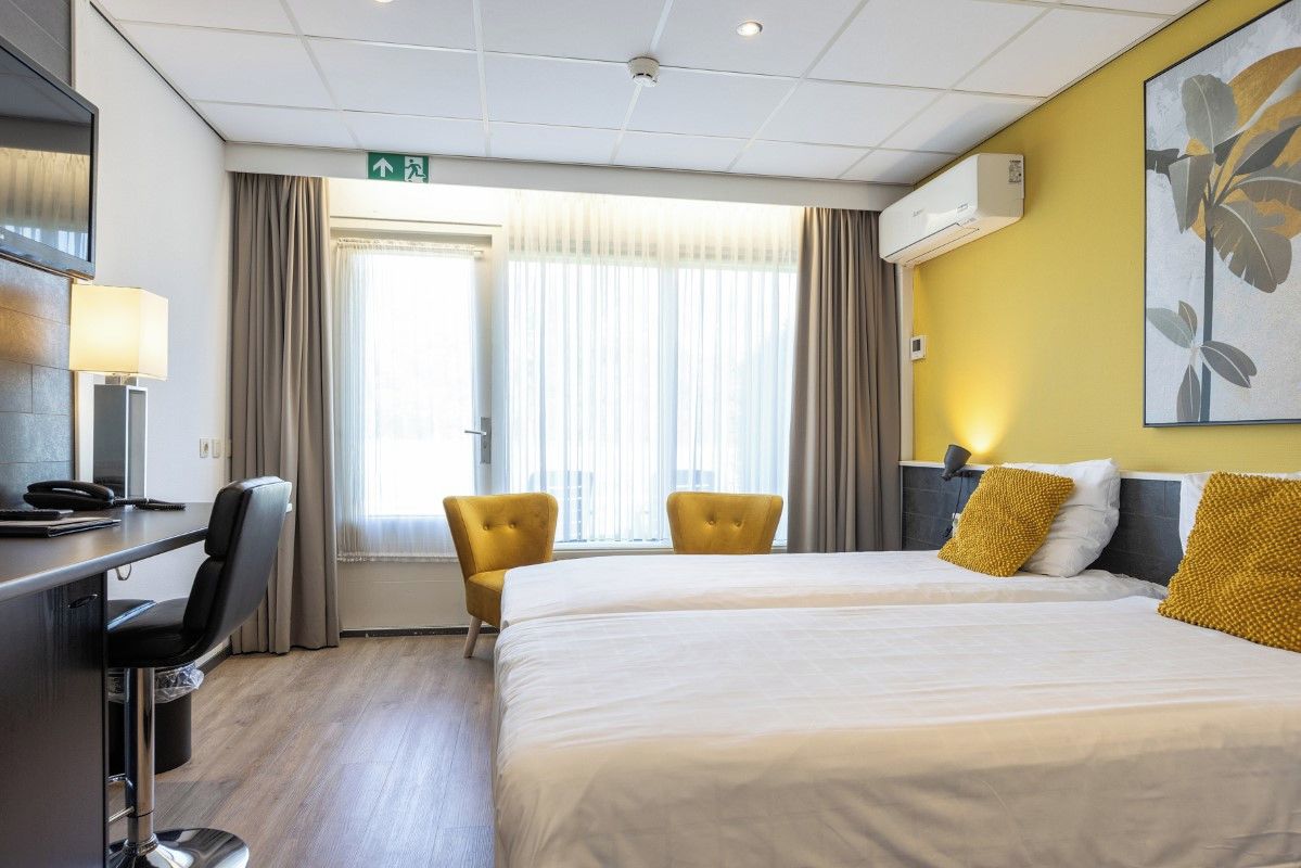 A hotel room with a bed, two chairs, a desk with a lamp, an air conditioner, and a window with curtains.