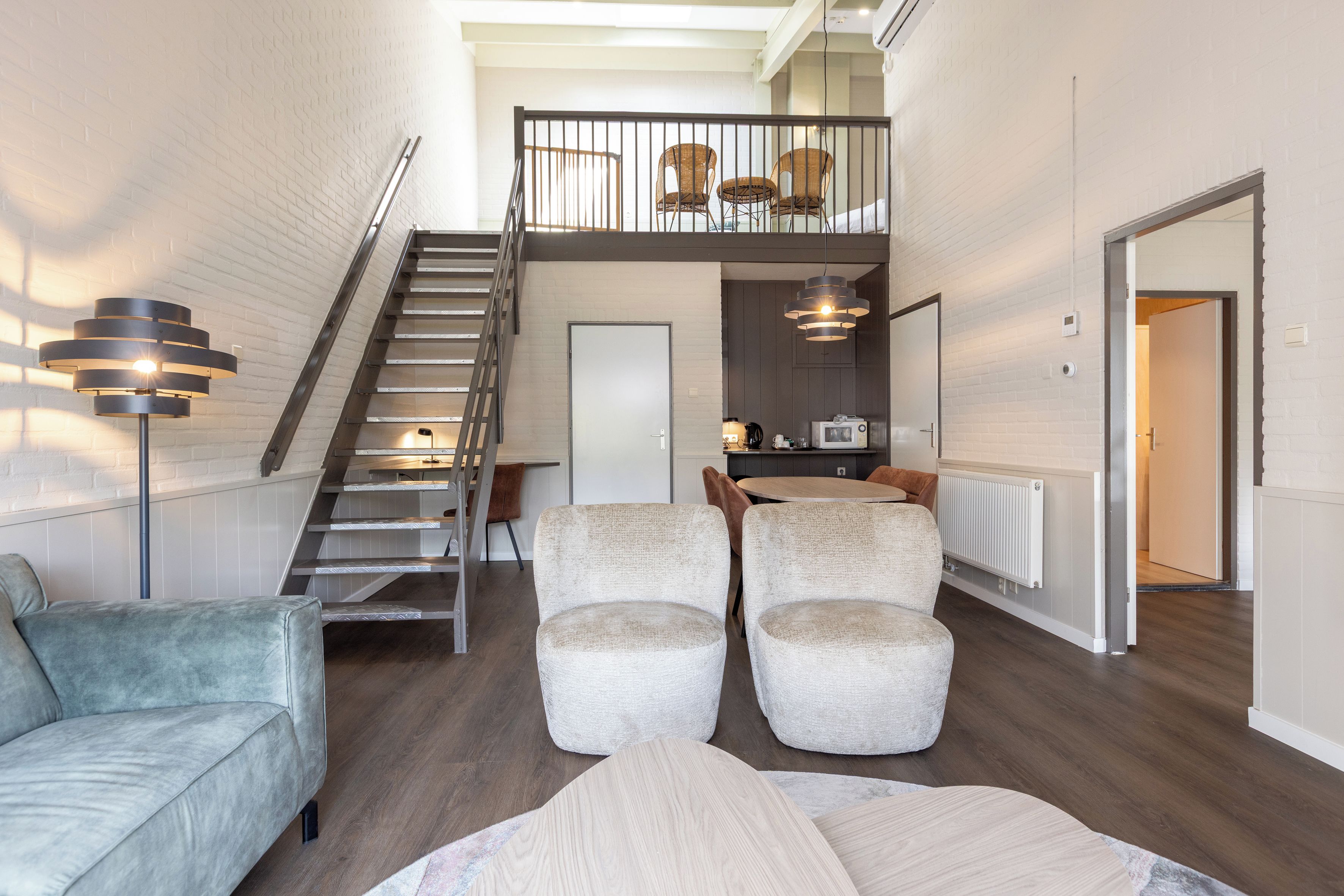 A spacious living area with a staircase leading to a loft, a sofa, armchairs, and a dining area with a table and chairs.